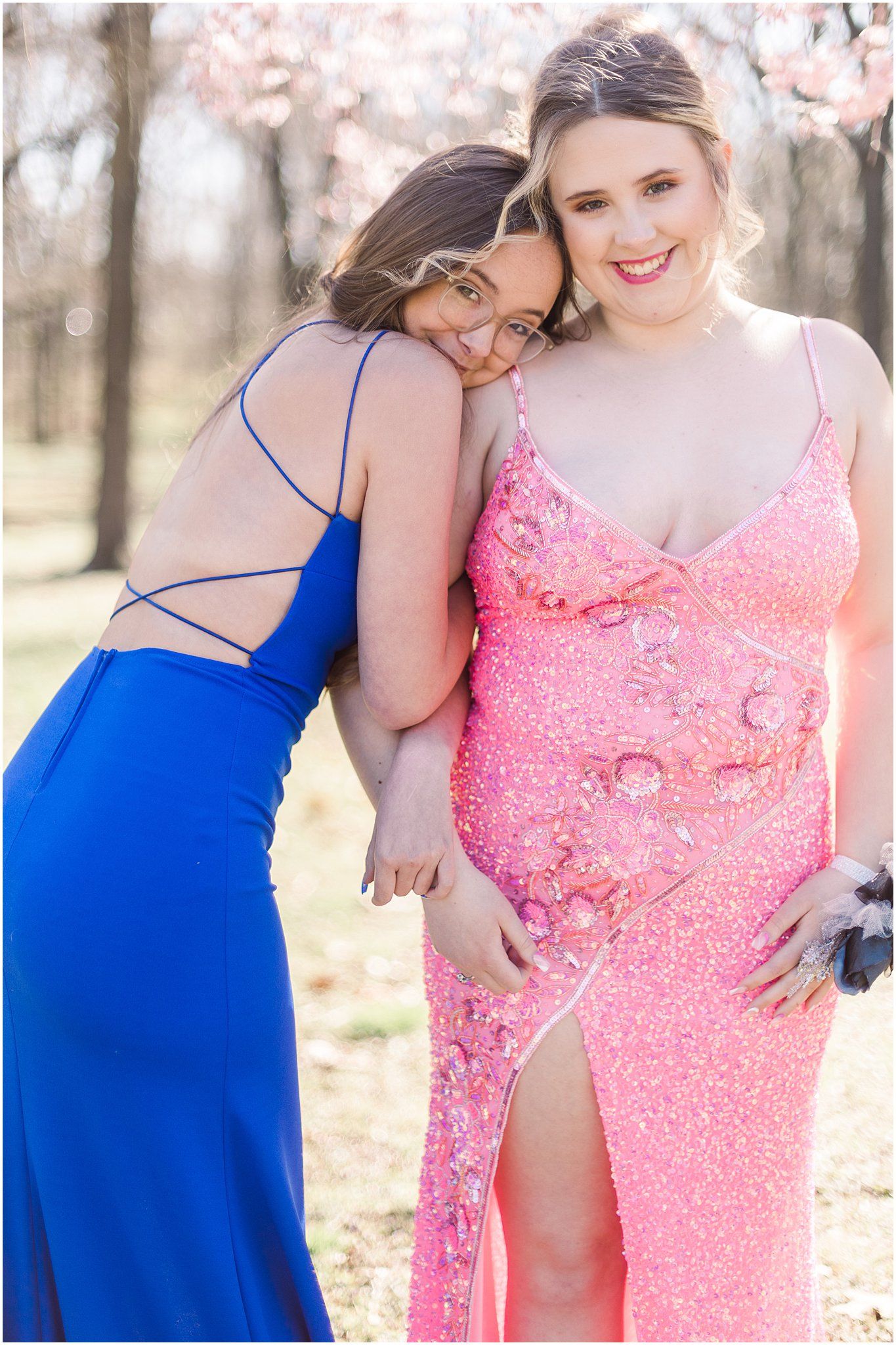 Two close friends share a tender moment in elegant prom dresses under spring blossoms.
