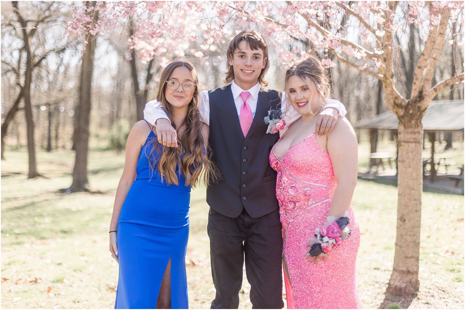 Three people in formal attire pose together under pink cherry blossom trees in springtime.