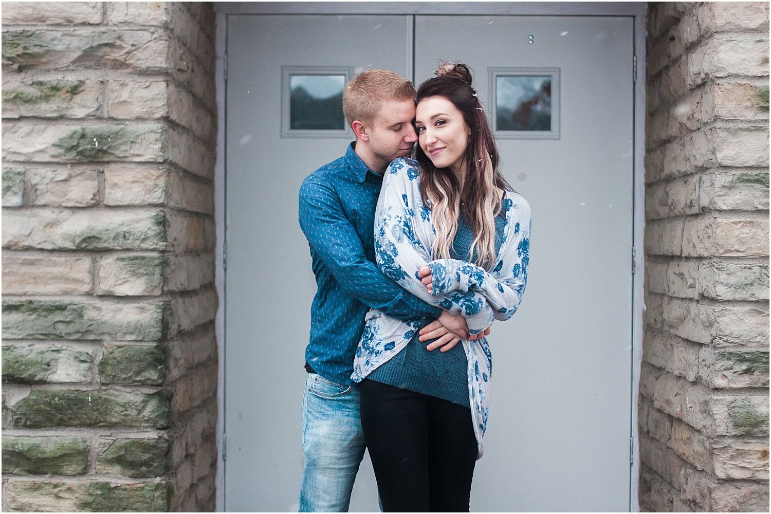 A couple poses intimately against a stone wall with a modern door backdrop.