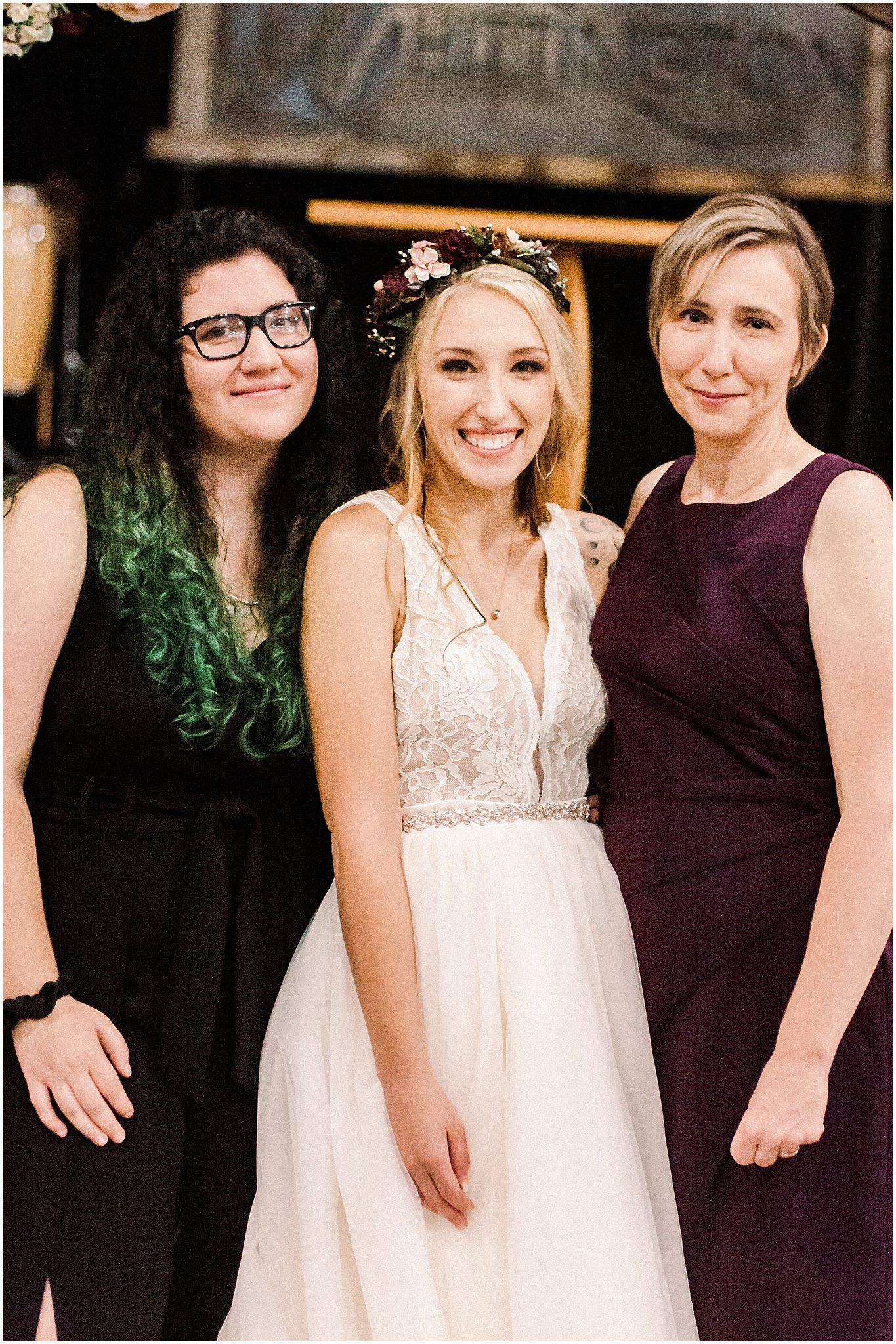 Three wedding attendees pose together under rustic wooden arch with floral decor.