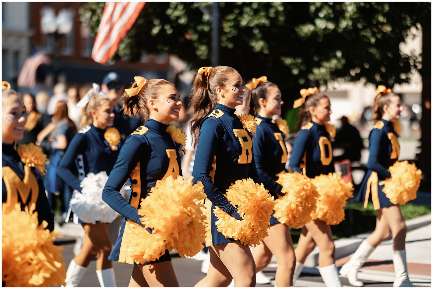 Line of cheerleaders in blue uniforms with yellow pom poms performing at outdoor event.