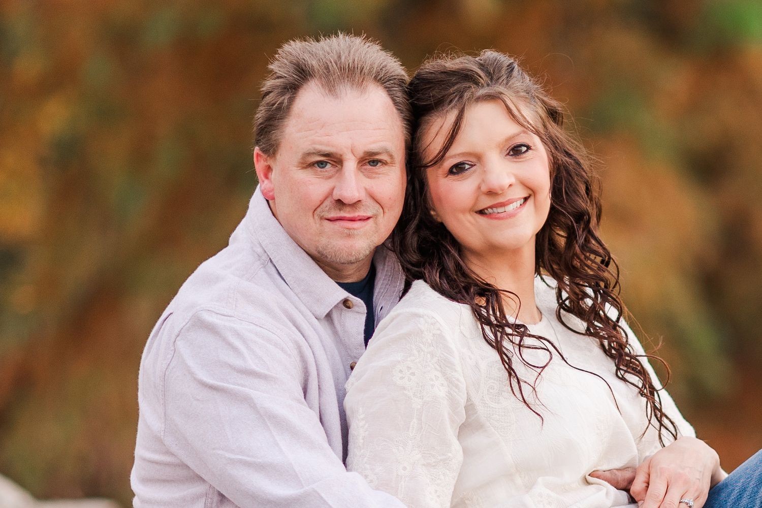 Portrait of couple in light colored sweaters against blurred fall foliage.