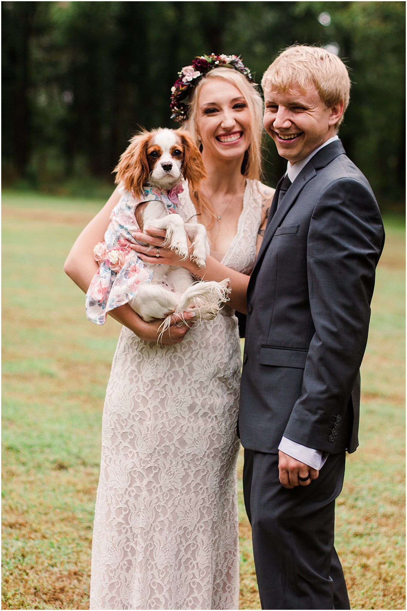 Happy couple poses with their Cavalier King Charles Spaniel at outdoor wedding.