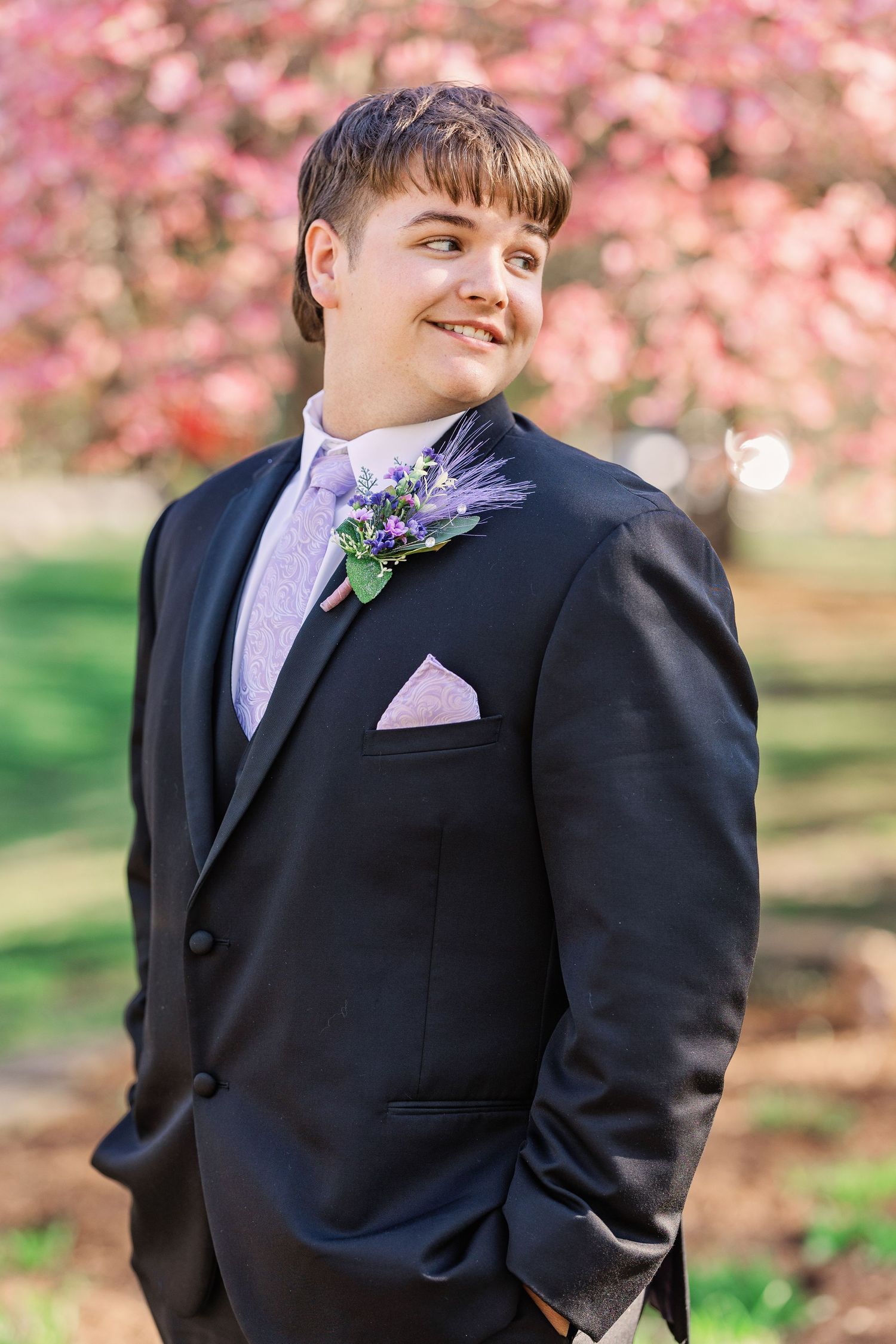 Young person in black tuxedo with purple tie and boutonniere poses under cherry blossoms.