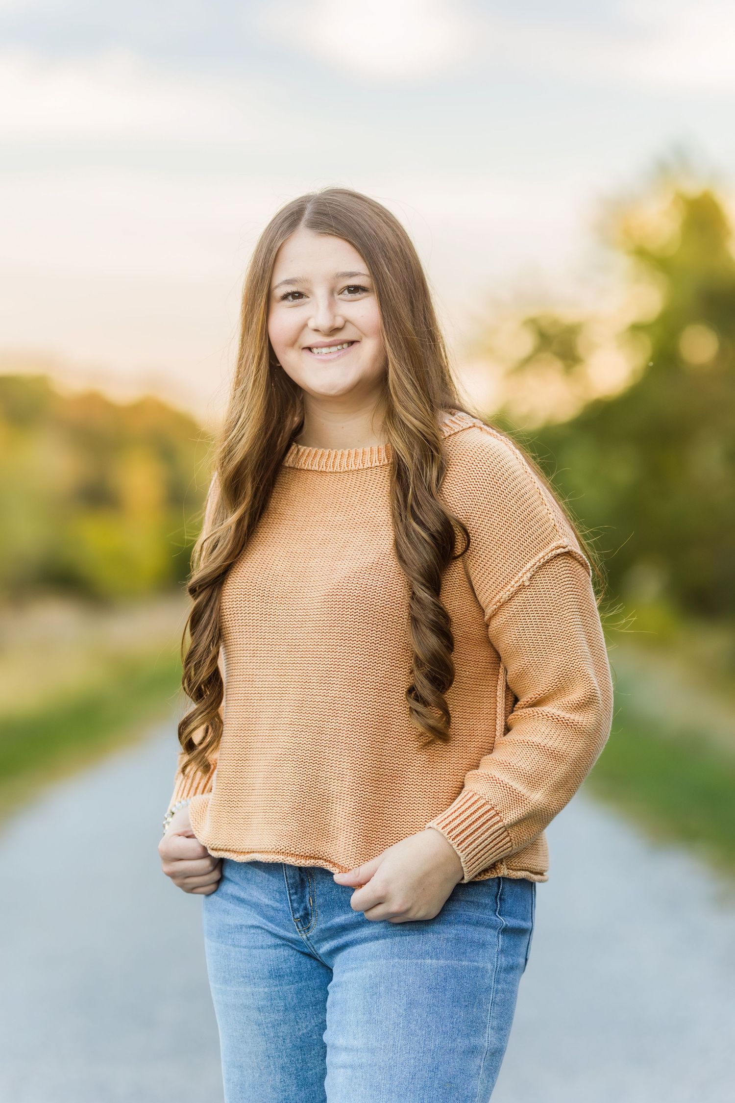A person in a tan sweater and jeans poses outdoors on a rural path during sunset.