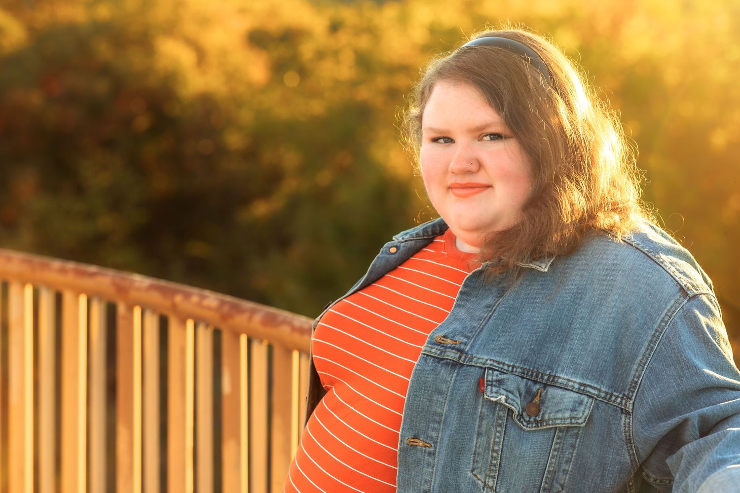 Person in orange top and denim jacket poses against autumn sunset backdrop.