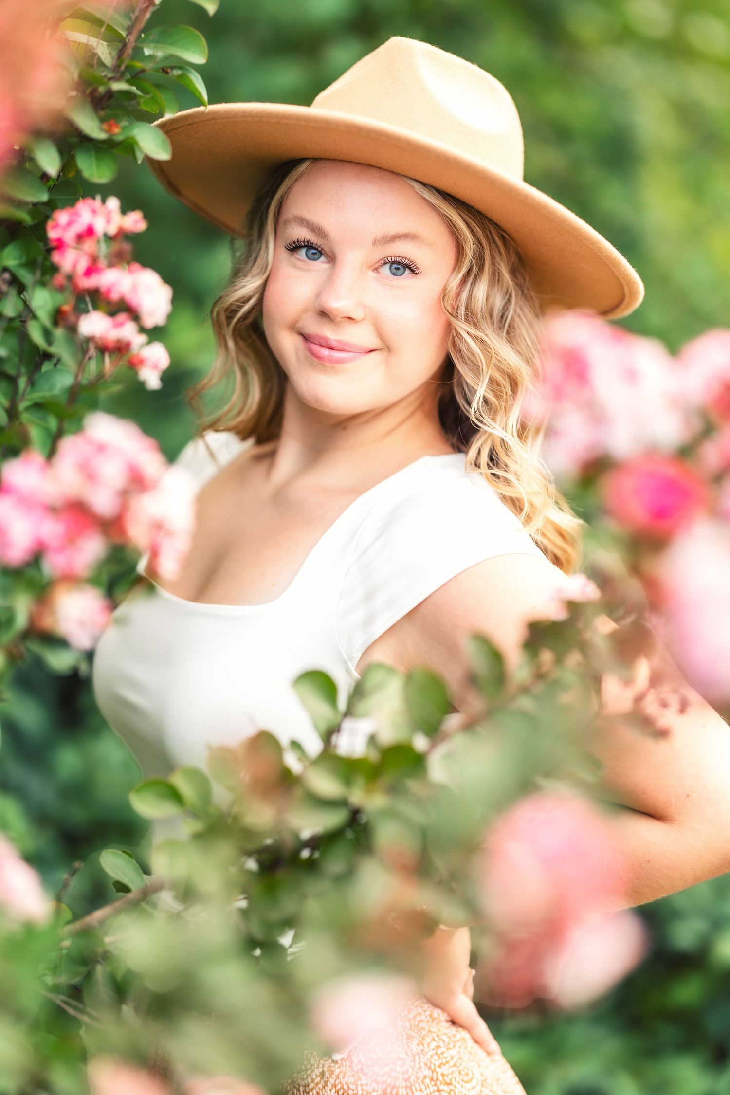 A romantic portrait among pink roses wearing a white top and tan hat.