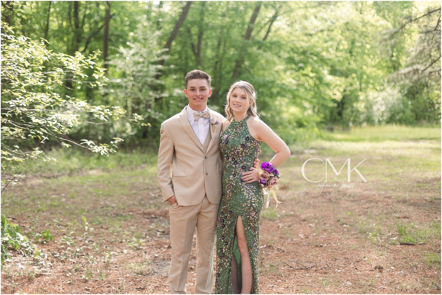 A formal couple poses together on a wooded path, wearing a tan suit and sequined green dress.