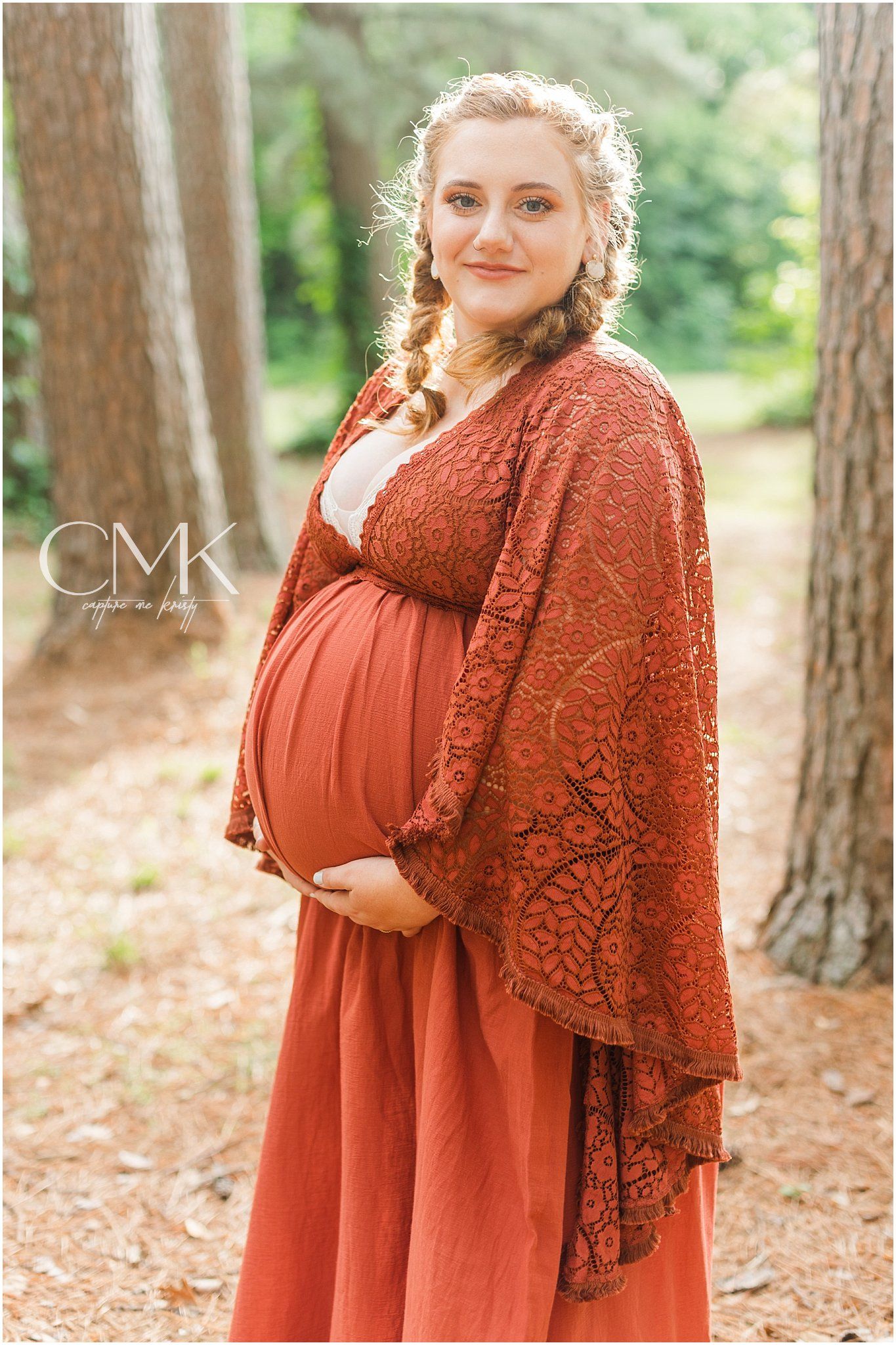Glowing mother-to-be in flowing rust dress poses among pine trees.