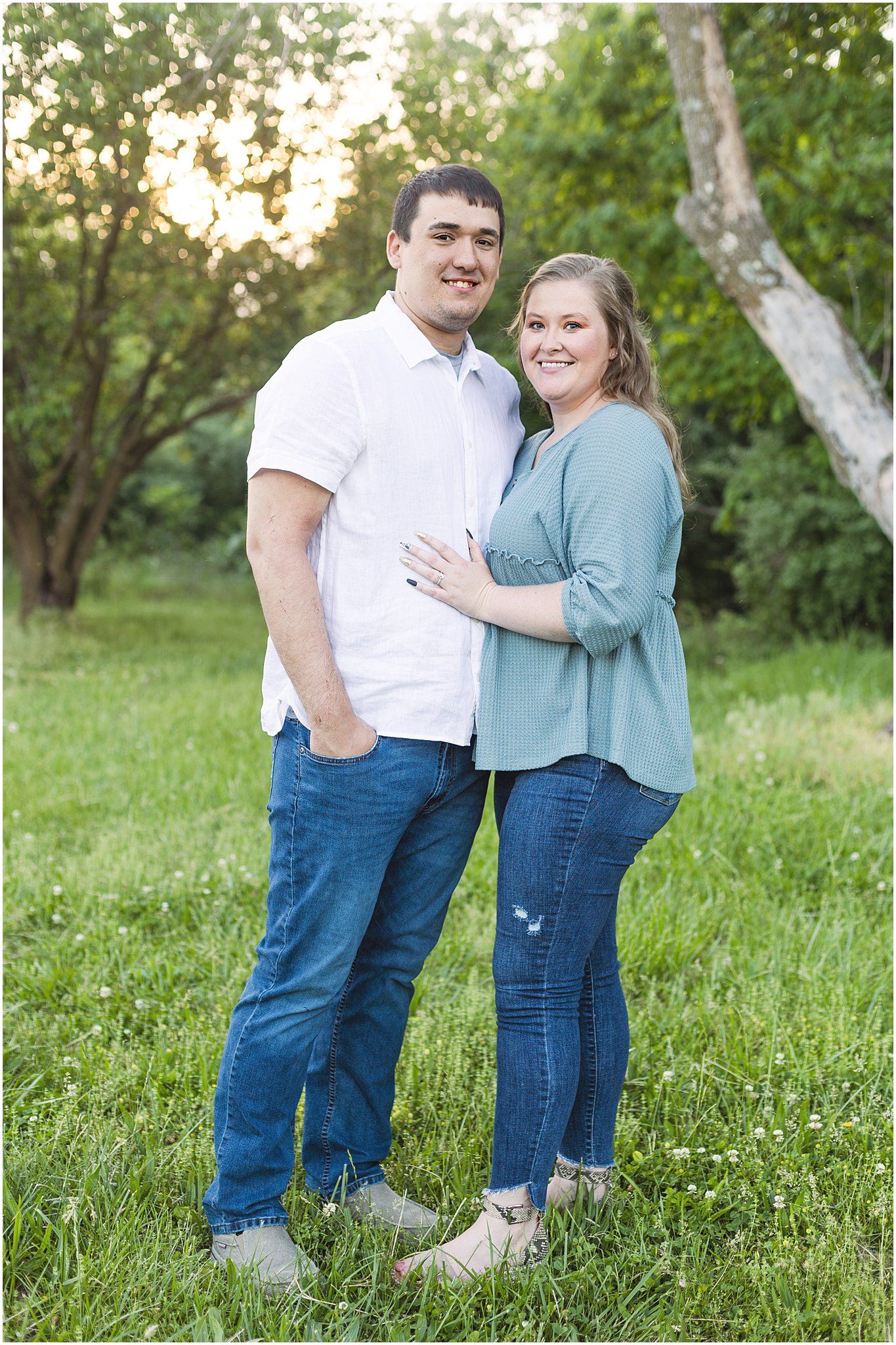 A couple poses together on grass with trees and sunset in background wearing casual blue jeans and white and blue tops.