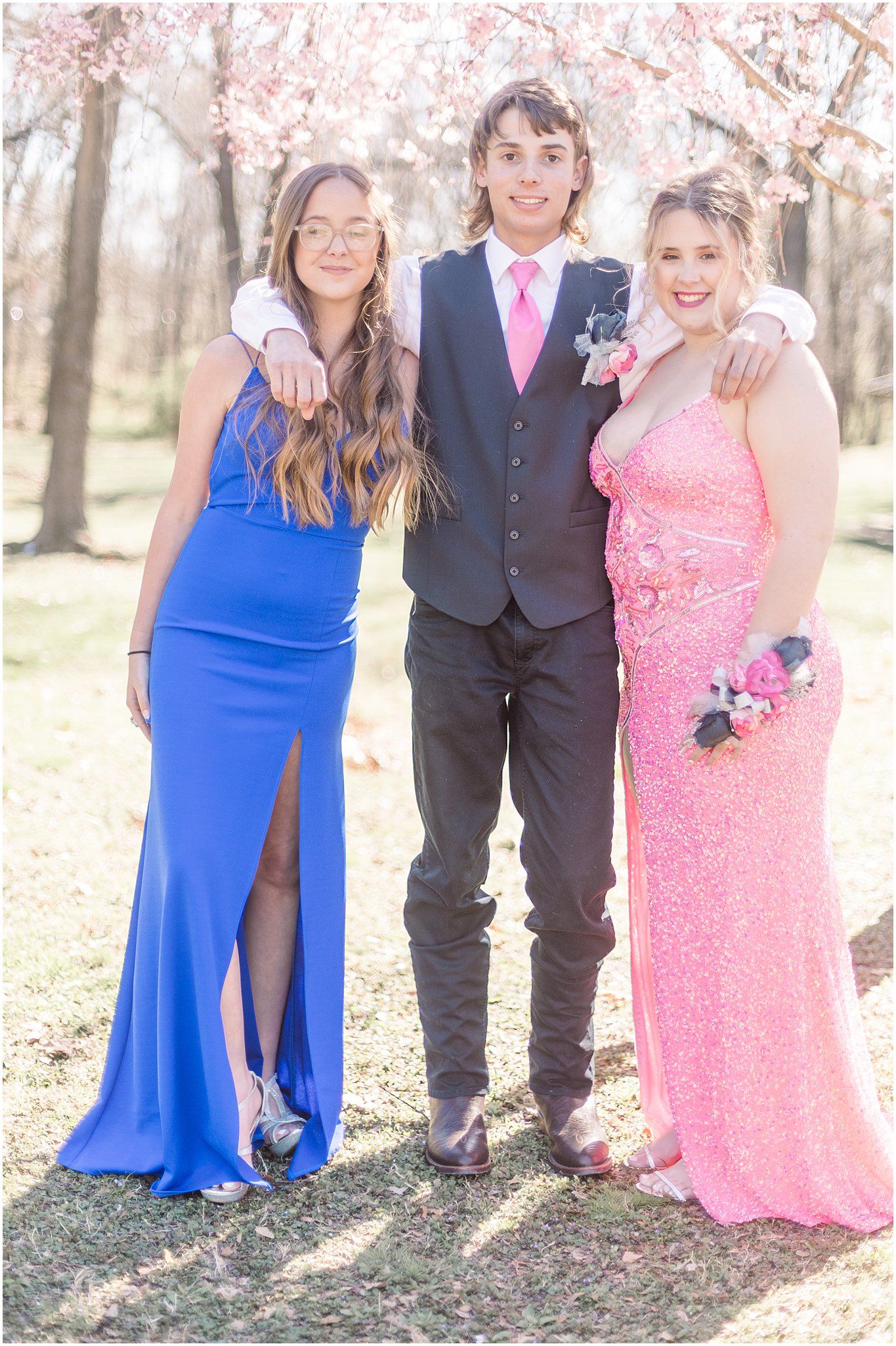 Friends pose in formal wear including a blue gown, black suit, and pink sequined dress under spring blossoms.