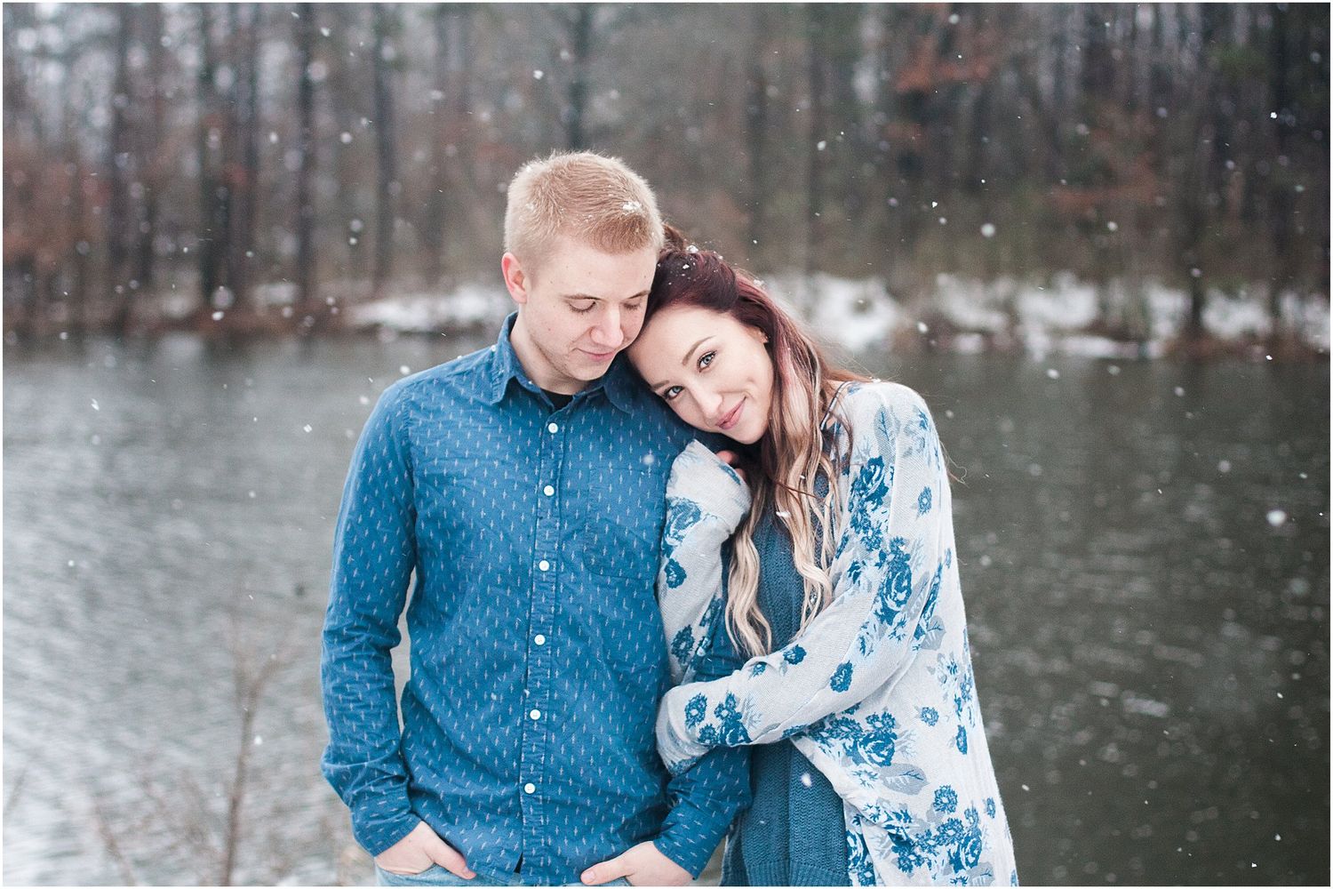 A romantic winter portrait of a couple embracing by a lake while snow falls around them.