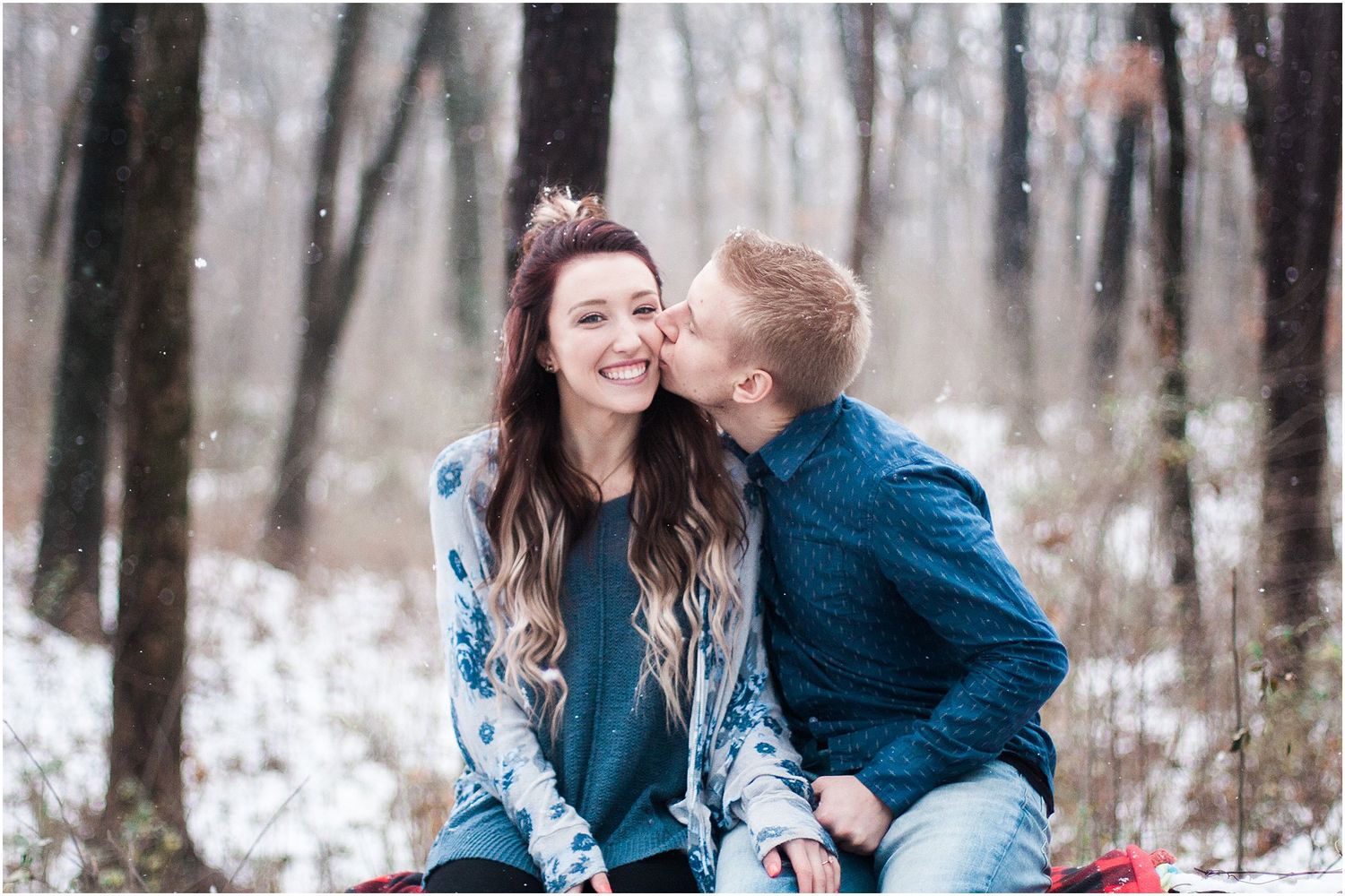 A romantic winter moment captured between a couple sitting on a red plaid blanket in the snow.