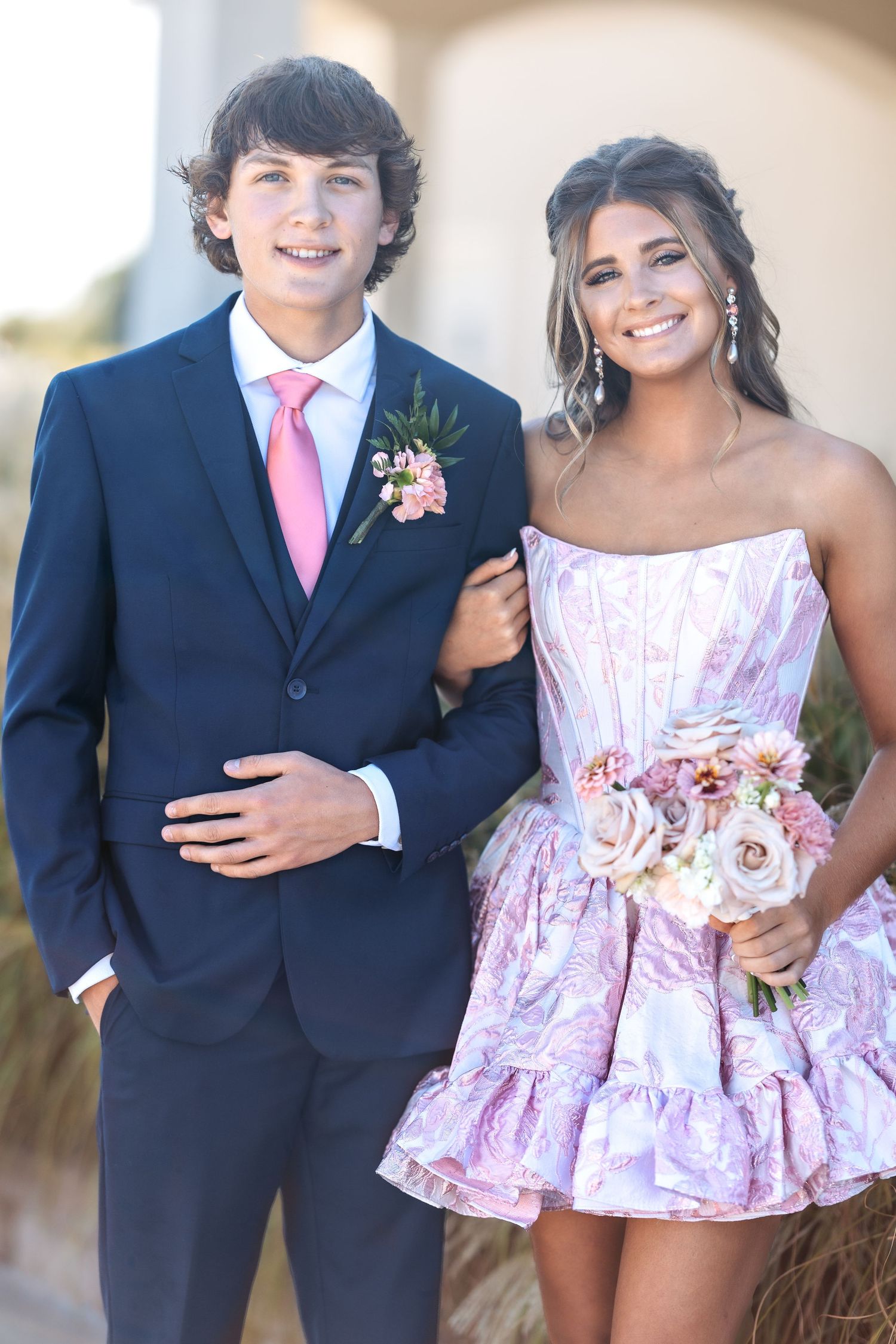 Formal pair in navy suit and pink dress pose together against light background.