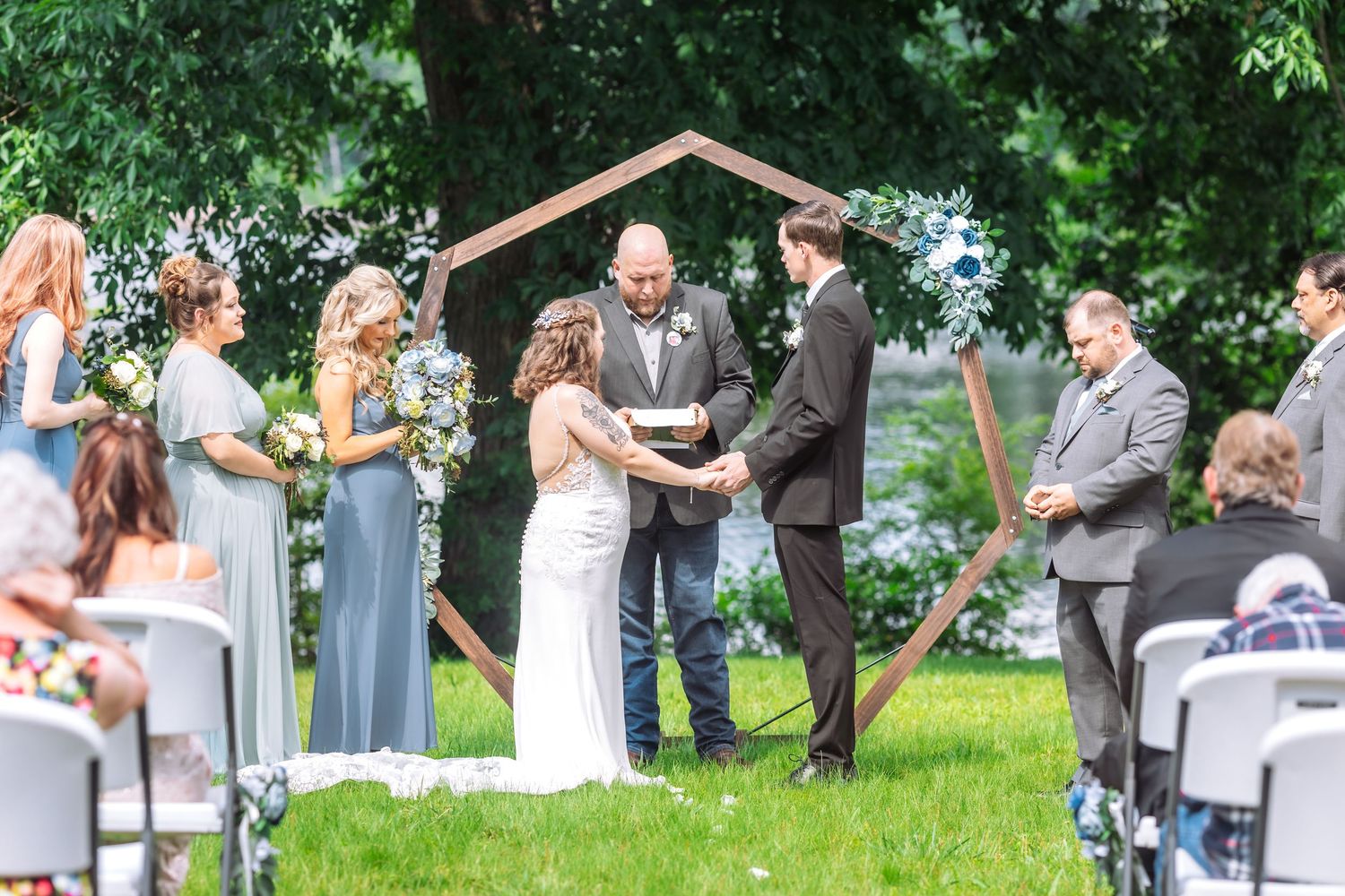 Wedding ceremony in progress under wooden arch with bridesmaids in blue dresses and guests seated.