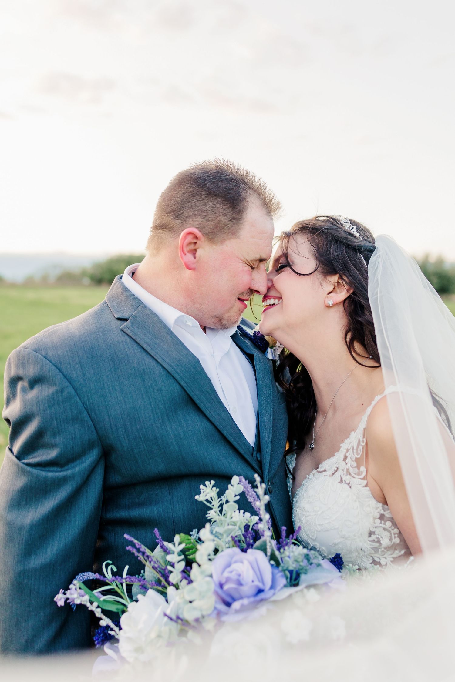 Romantic outdoor wedding portrait with purple bouquet and flowing veil.