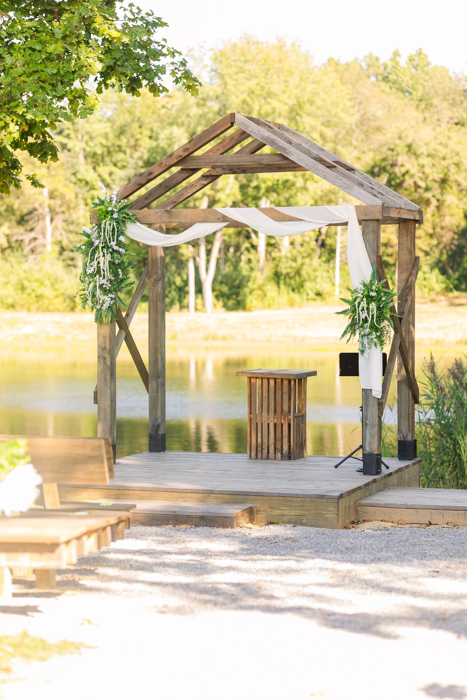 Rustic wooden pergola with white draping overlooks a serene lake in a natural outdoor setting.