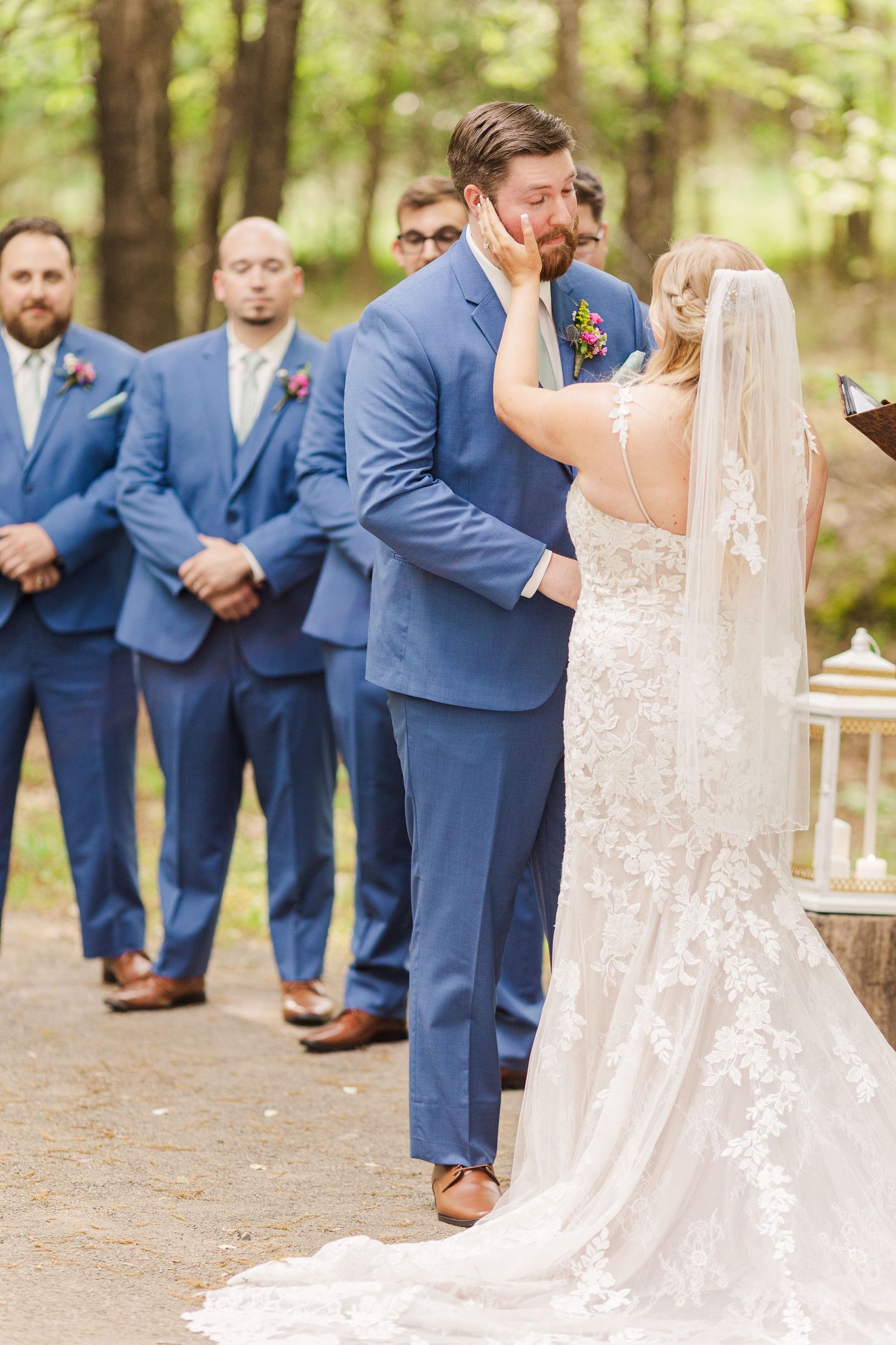 Groomsmen in navy suits line up outdoors while a bride's lace dress train extends behind her.