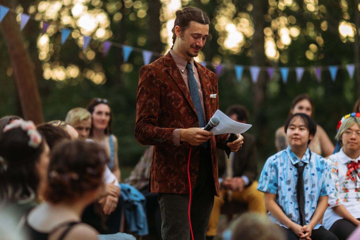 A speaker in a burgundy jacket addresses wedding guests during an outdoor forest celebration.