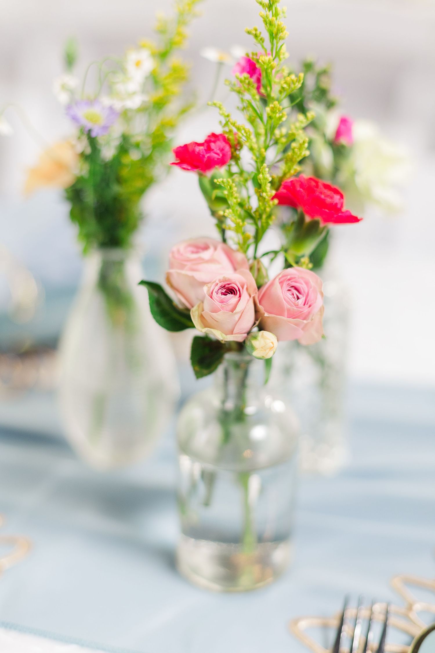 Simple floral centerpieces with pink roses and greenery in glass vases line a table.