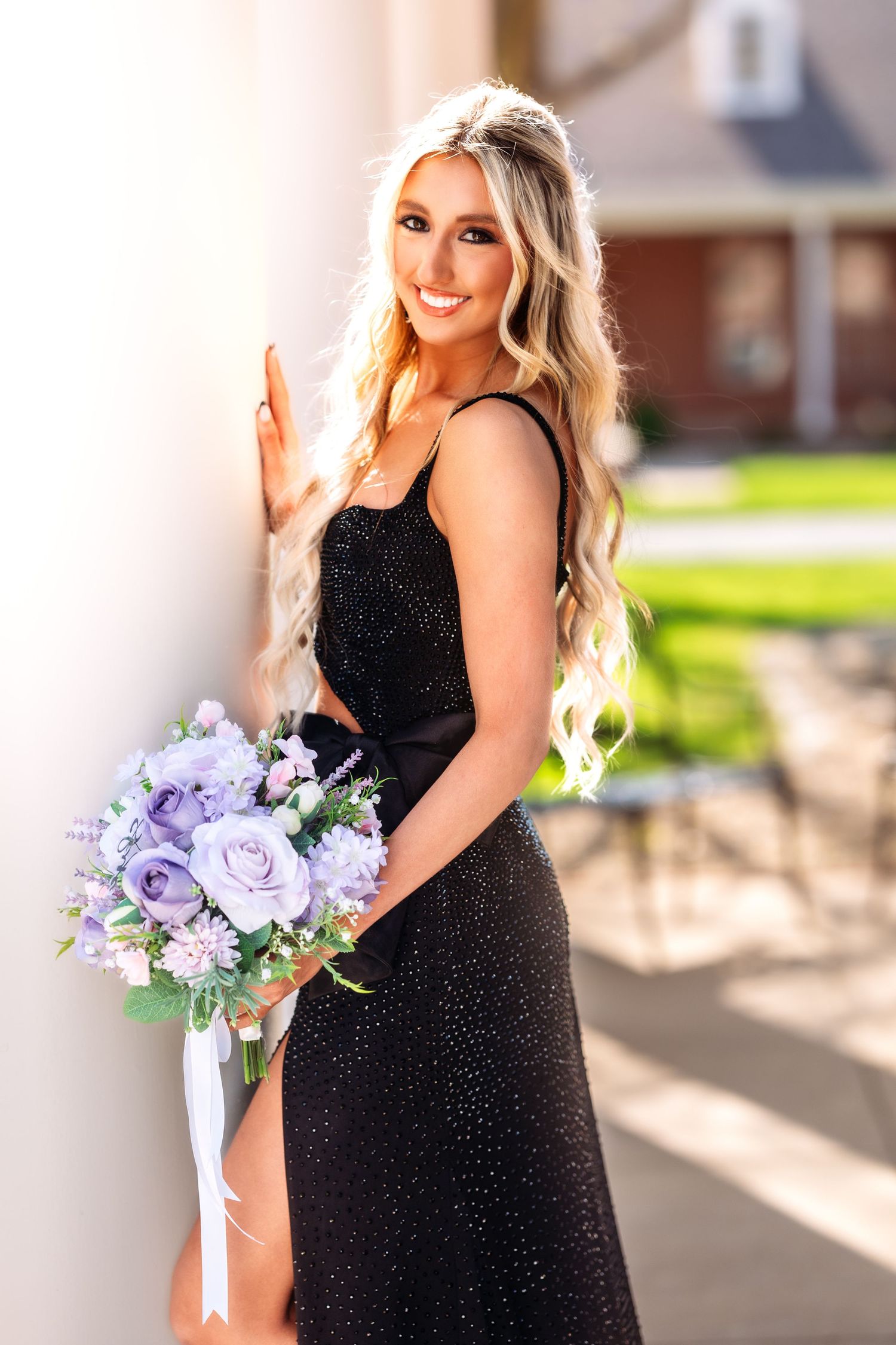 Woman in sparkling black formal dress holds purple and white floral bouquet while posing outdoors in sunlight.