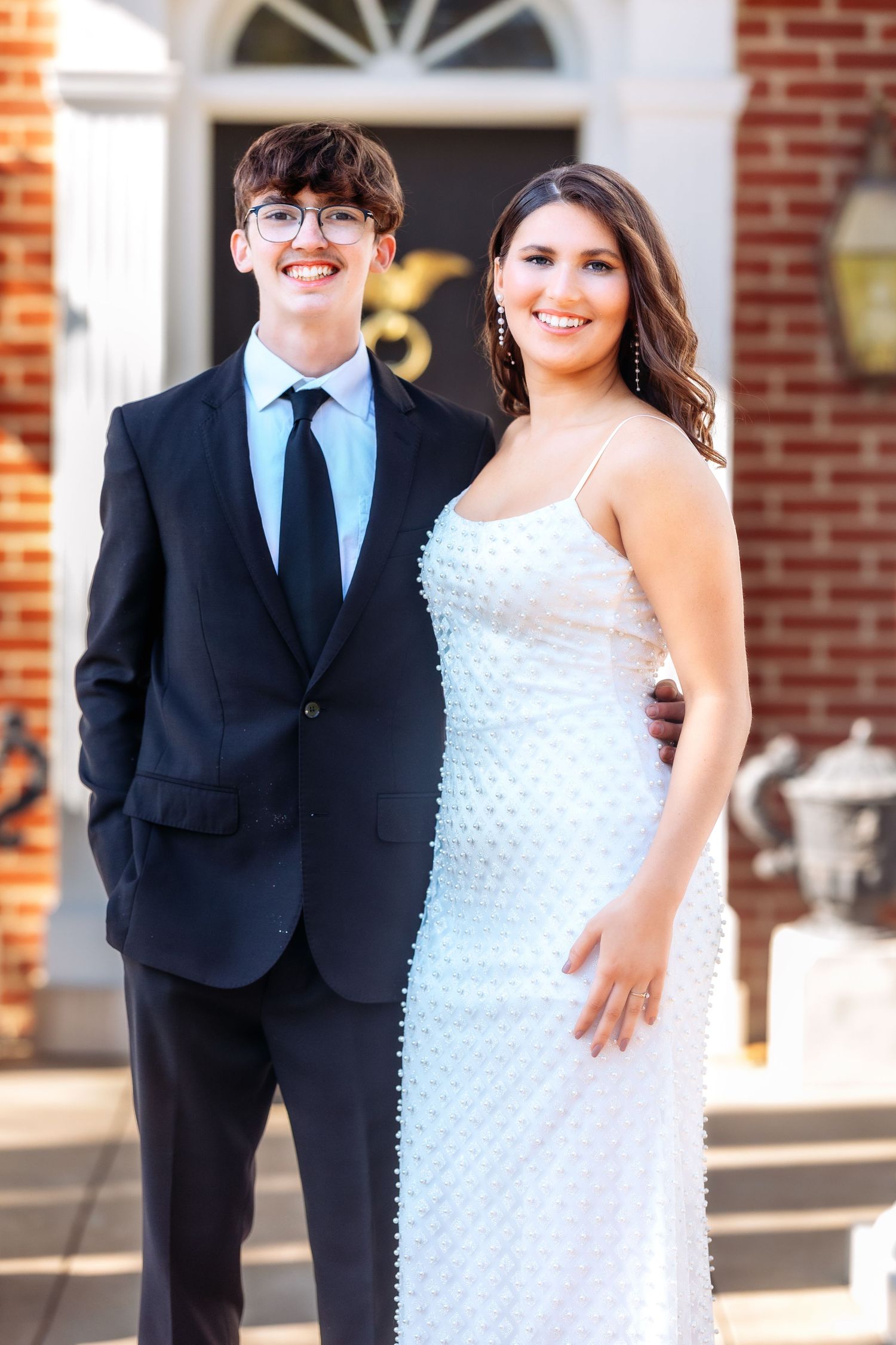A couple poses together in formal attire, with the woman wearing a white textured dress and the man in a dark suit.