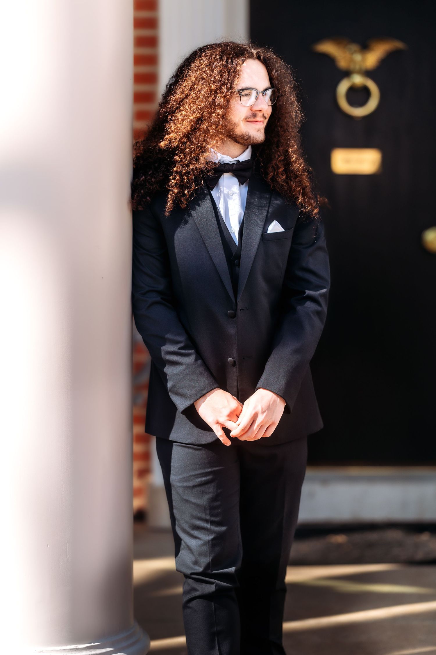 Person with long curly hair wearing a black suit stands in elegant pose against architectural columns.