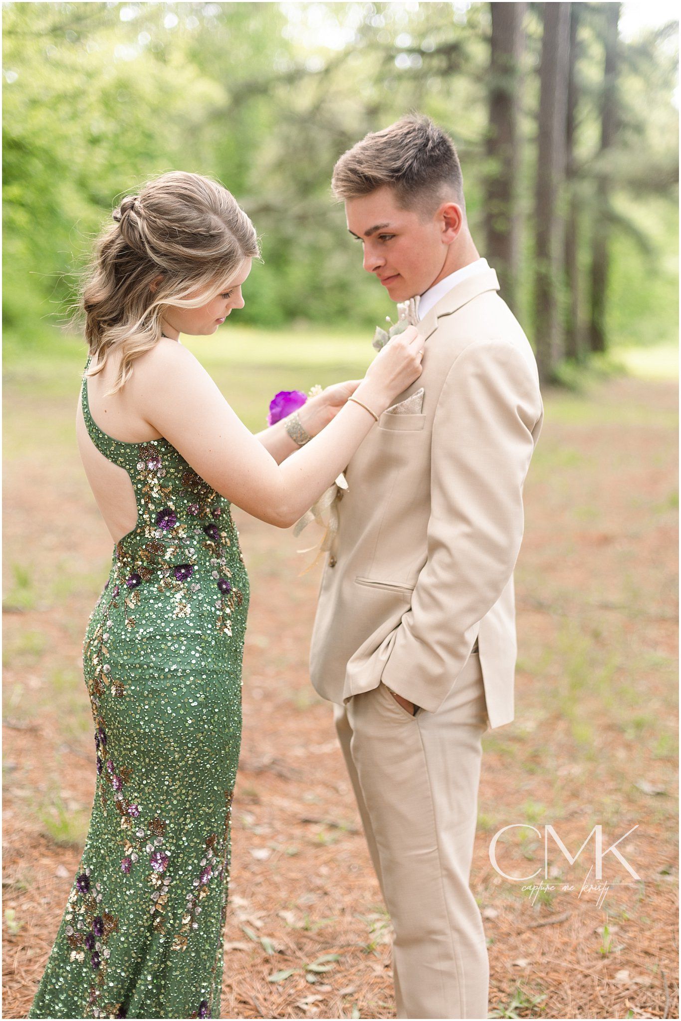 A couple in formal attire pins on corsages in a forest setting, she in a sparkly green dress and he in a tan suit.