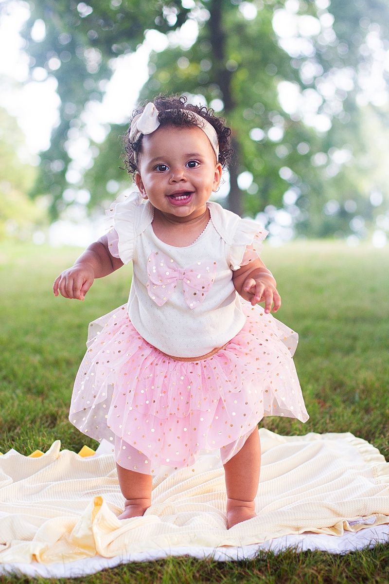 Baby wearing white and pink tulle dress with floral headband stands joyfully on blanket in grassy outdoor setting.
