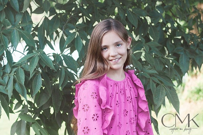 Portrait of young person in bright pink eyelet dress standing against green foliage backdrop.
