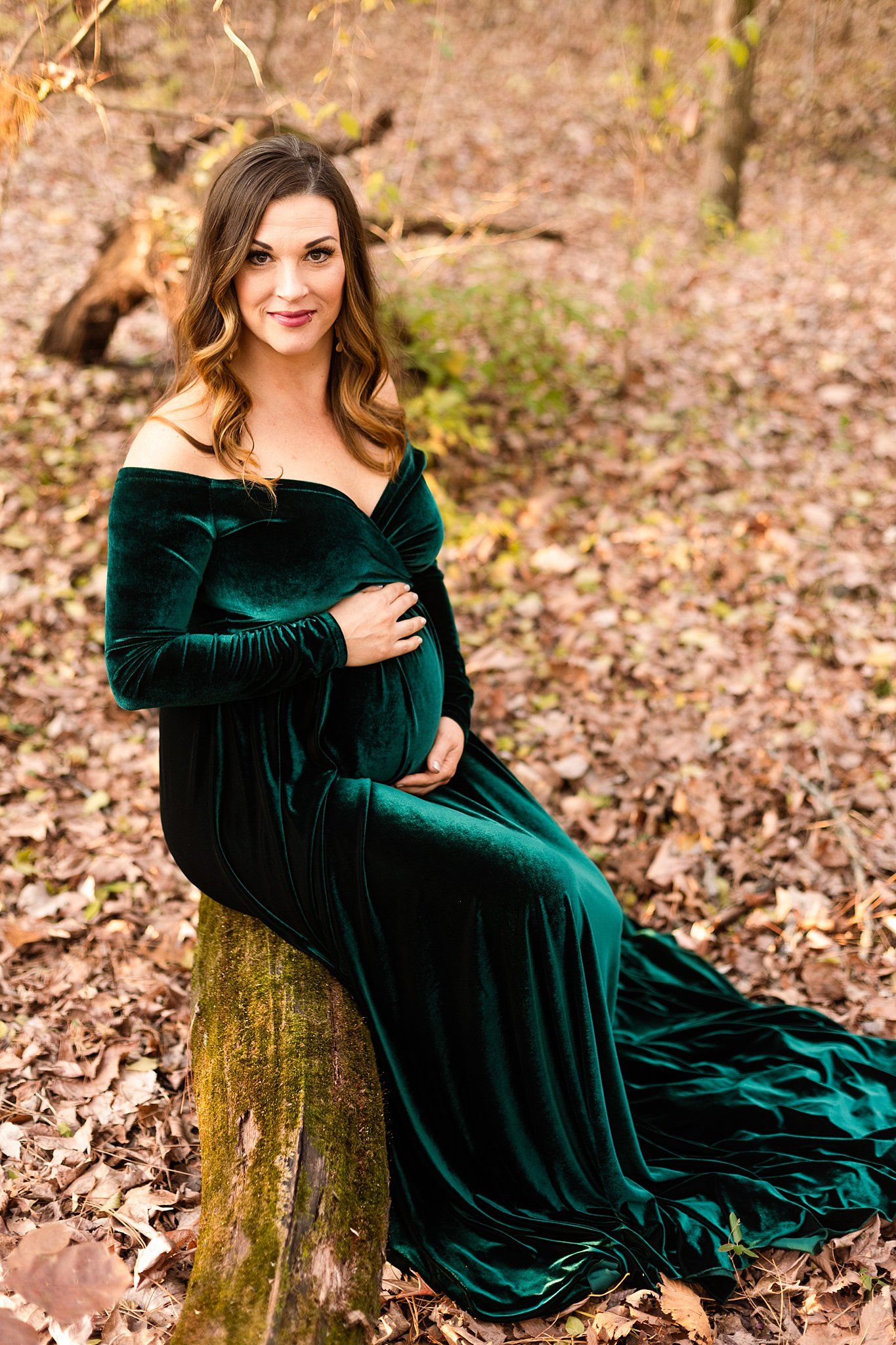 Smiling portrait in emerald velvet maternity gown seated on mossy log.