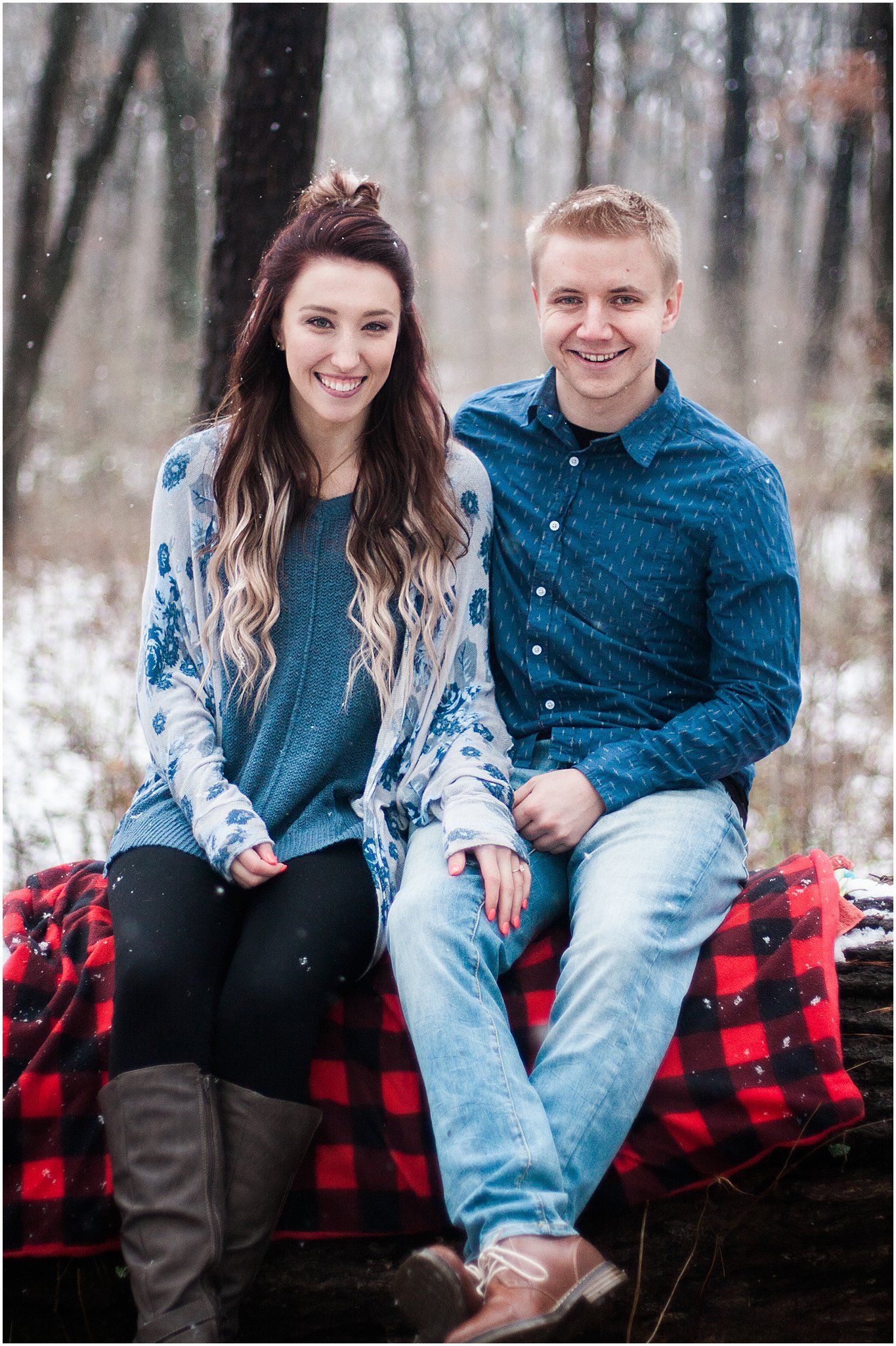 A couple sits on a red buffalo plaid blanket in a snowy winter forest setting wearing coordinating blue outfits.