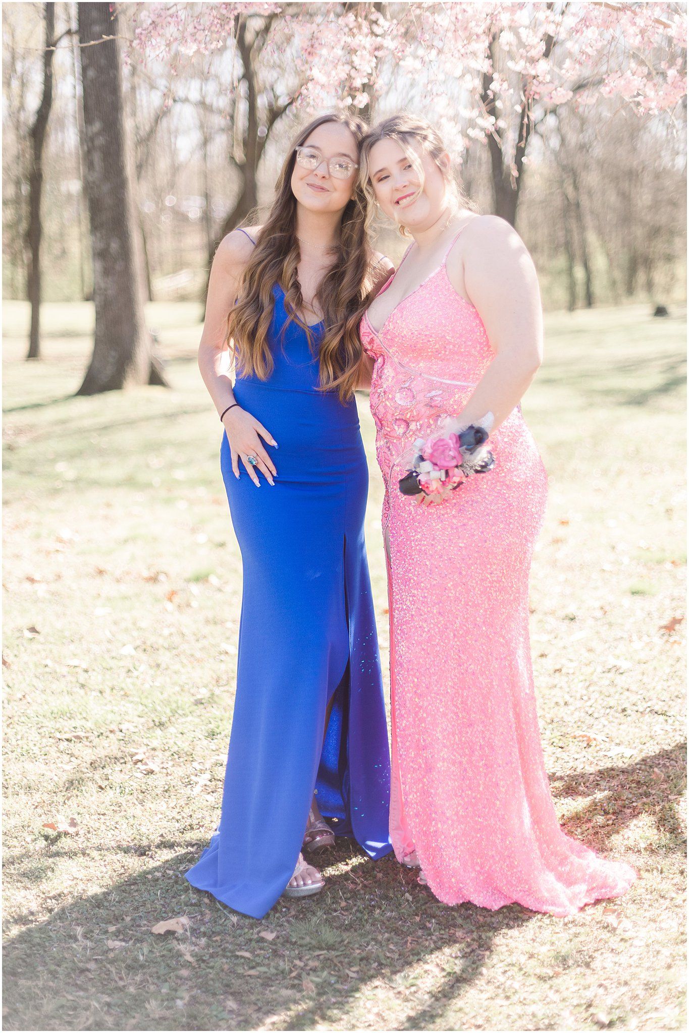 Two friends in formal gowns, one pink and one blue, pose under cherry blossoms.