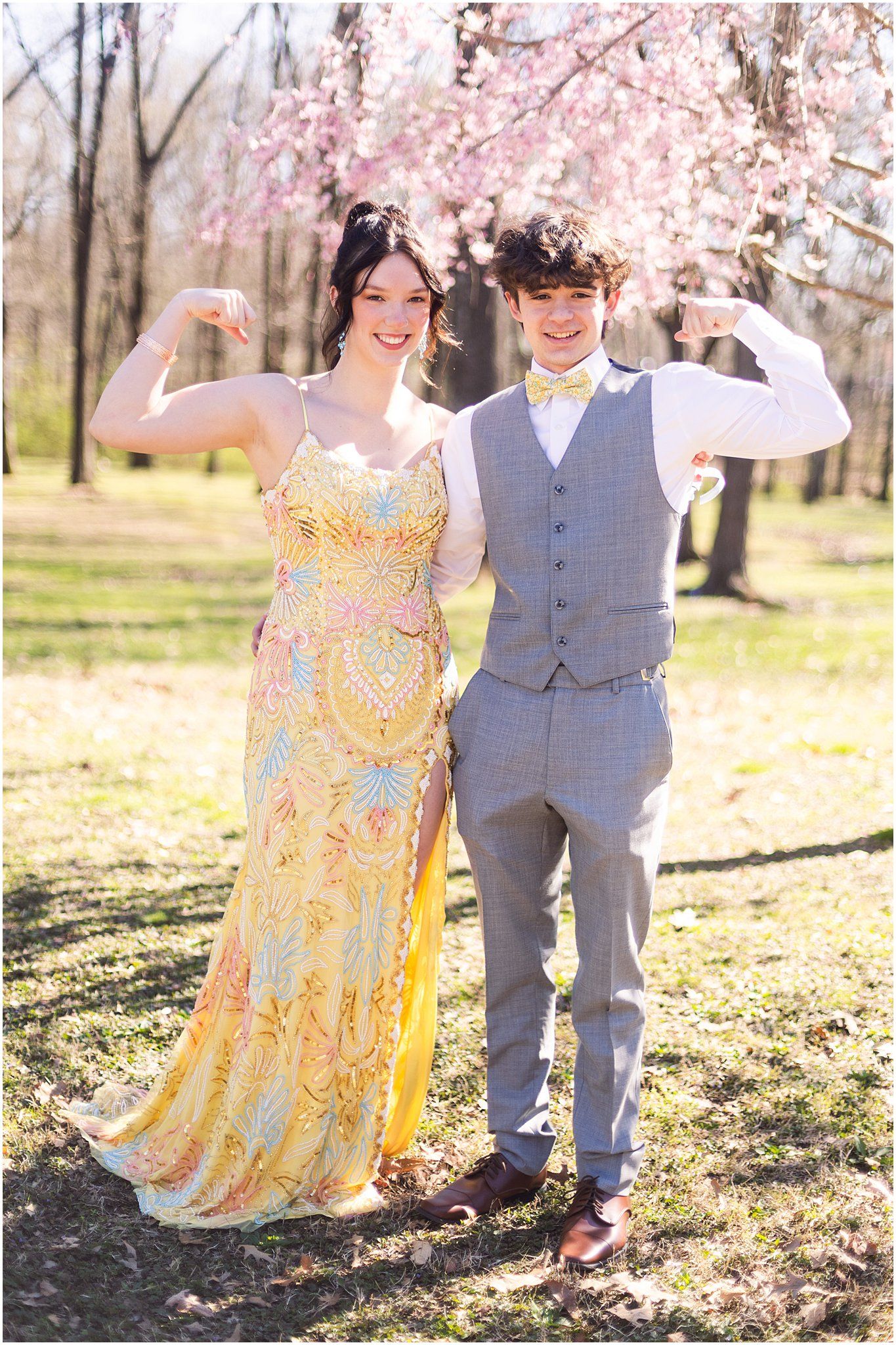 A couple poses outdoors in formal attire, with yellow dress and gray vest under blooming cherry blossoms.