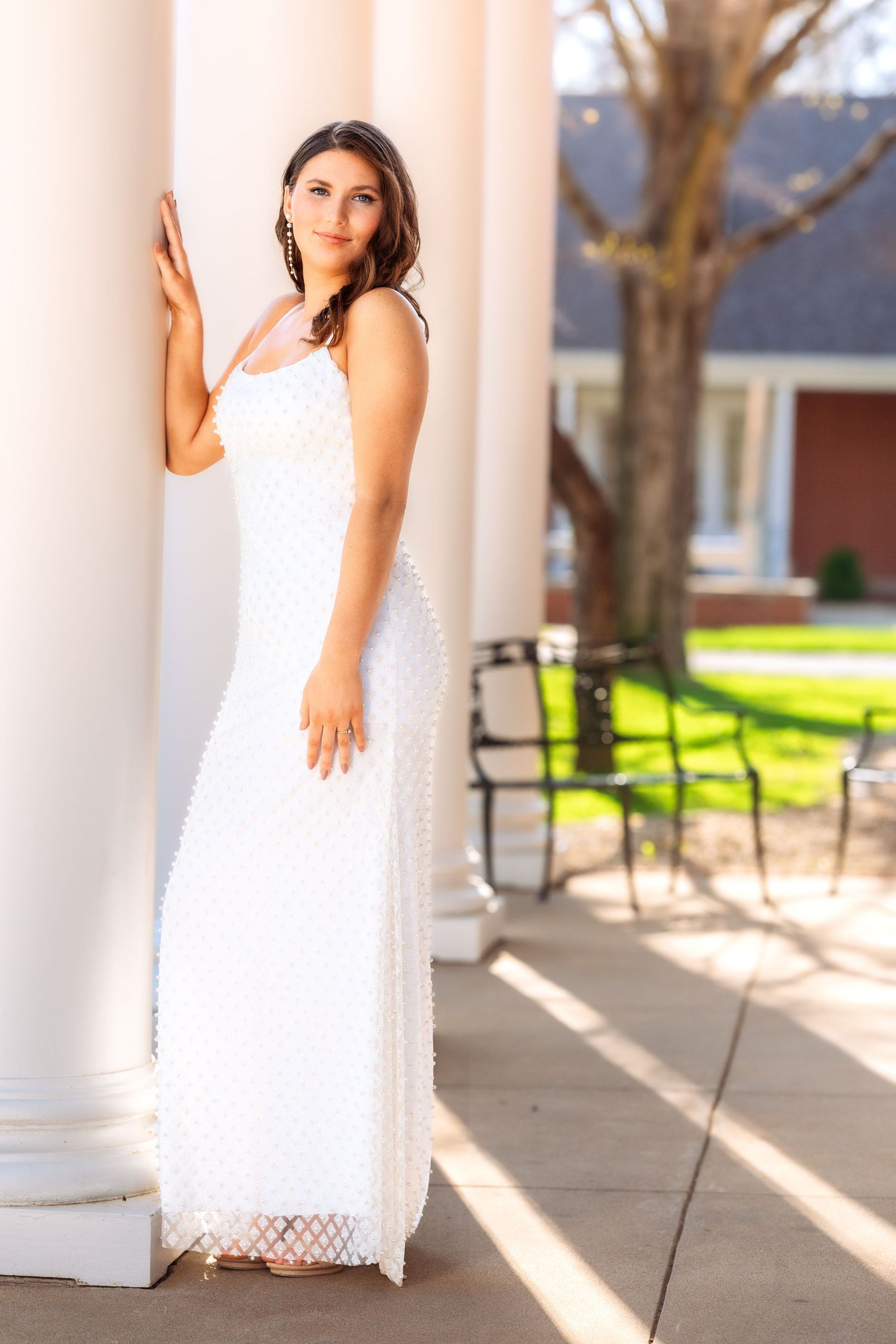 Woman in white lace dress poses elegantly on a sunlit porch with white columns and benches in background.