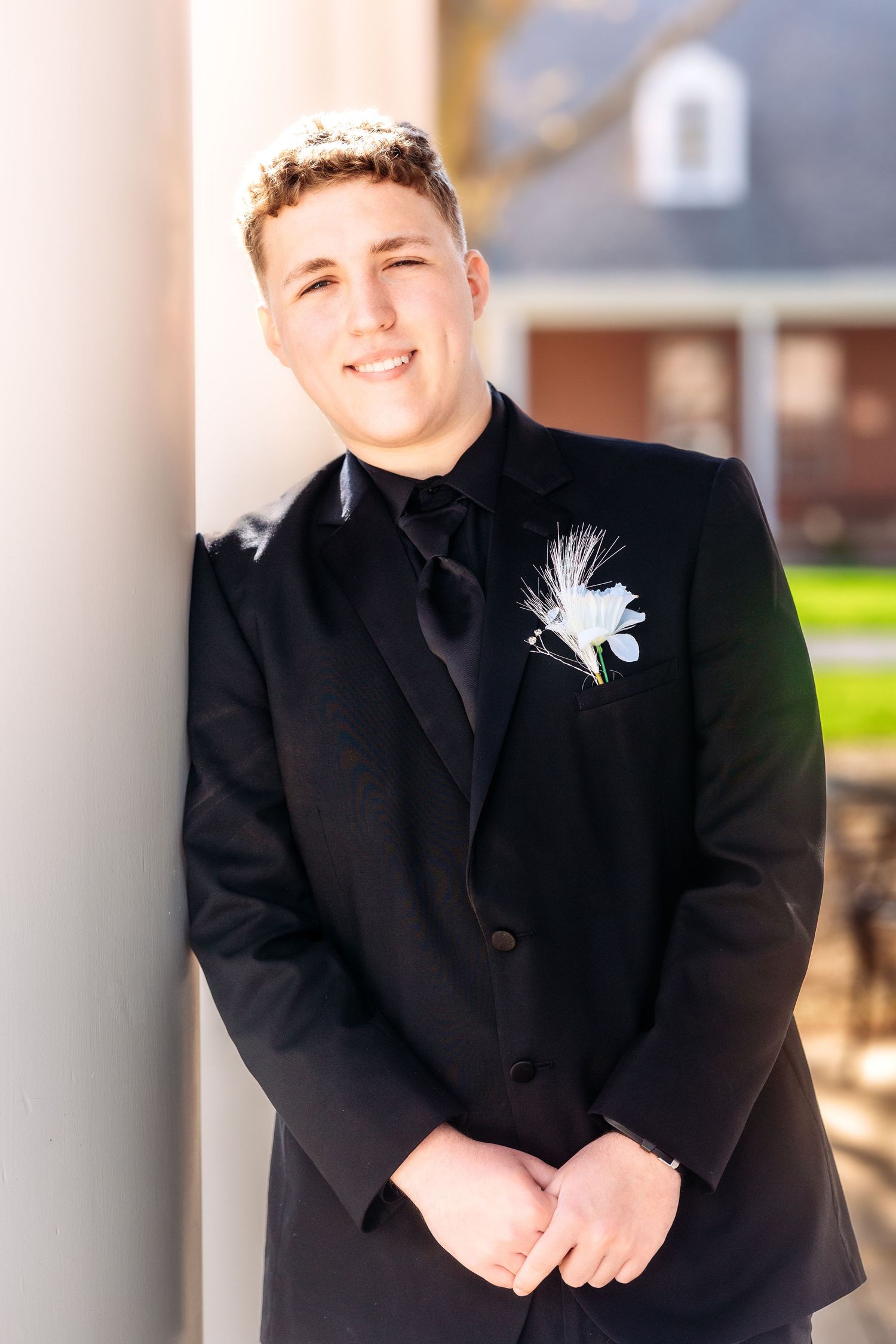 A person wearing a black formal suit with a white boutonniere smiles warmly in an outdoor setting.