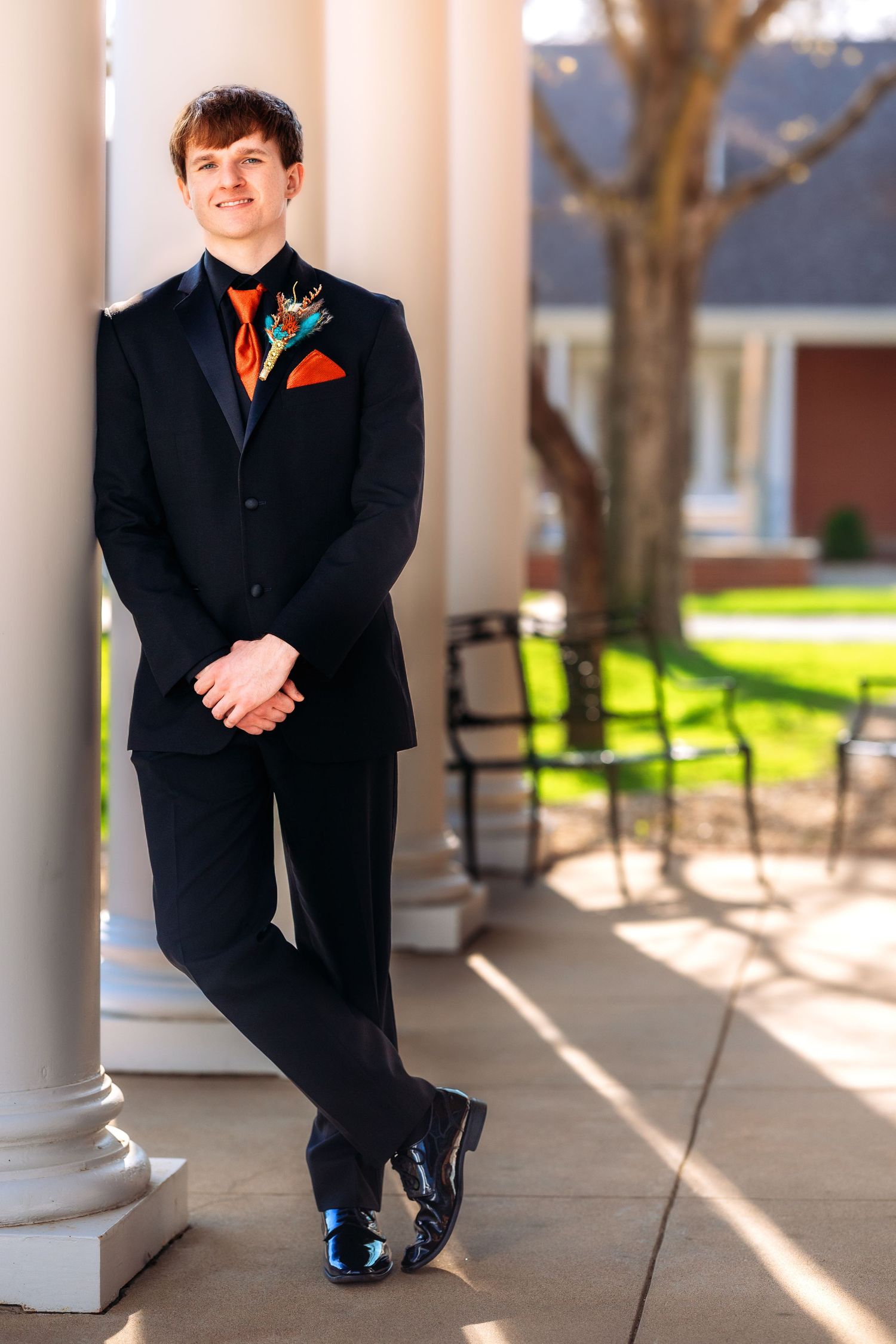 A person in a formal black suit with orange boutonniere leans against a white column on a sunny porch.