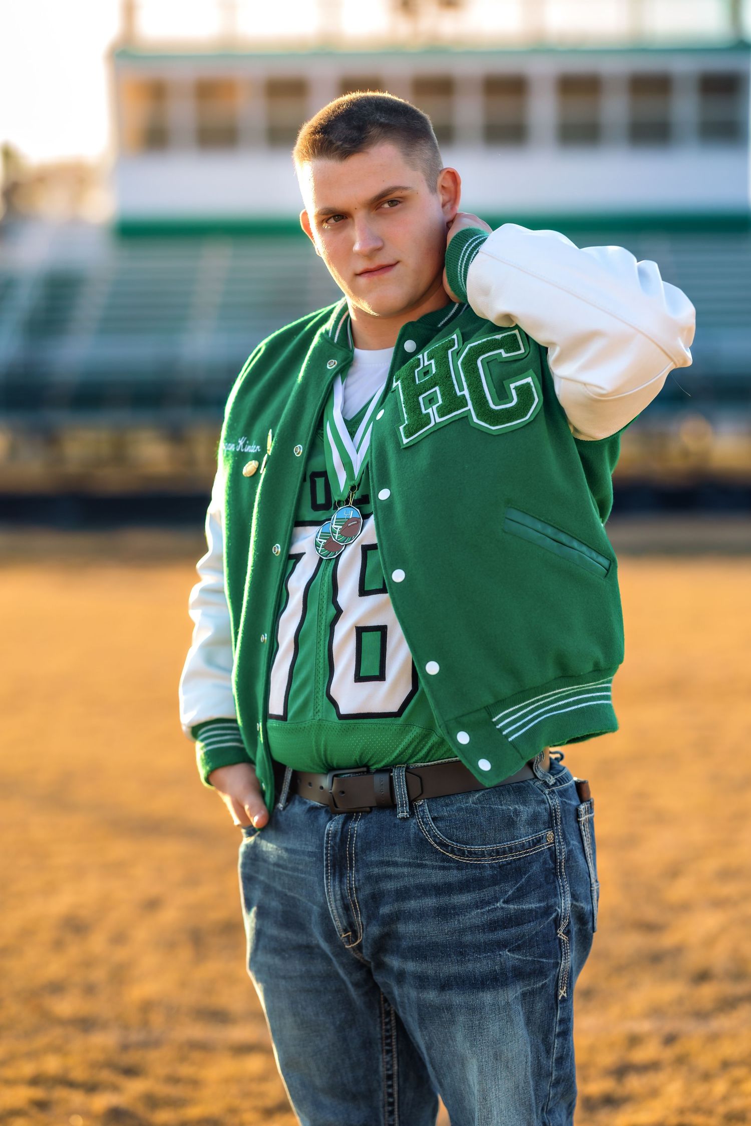 Athletic portrait in green varsity letterman jacket on sports field at sunset.