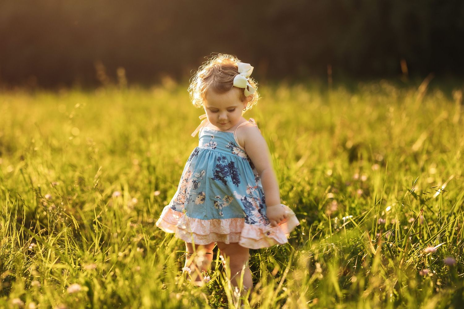 A toddler in a blue floral dress twirls in a golden sunlit meadow.