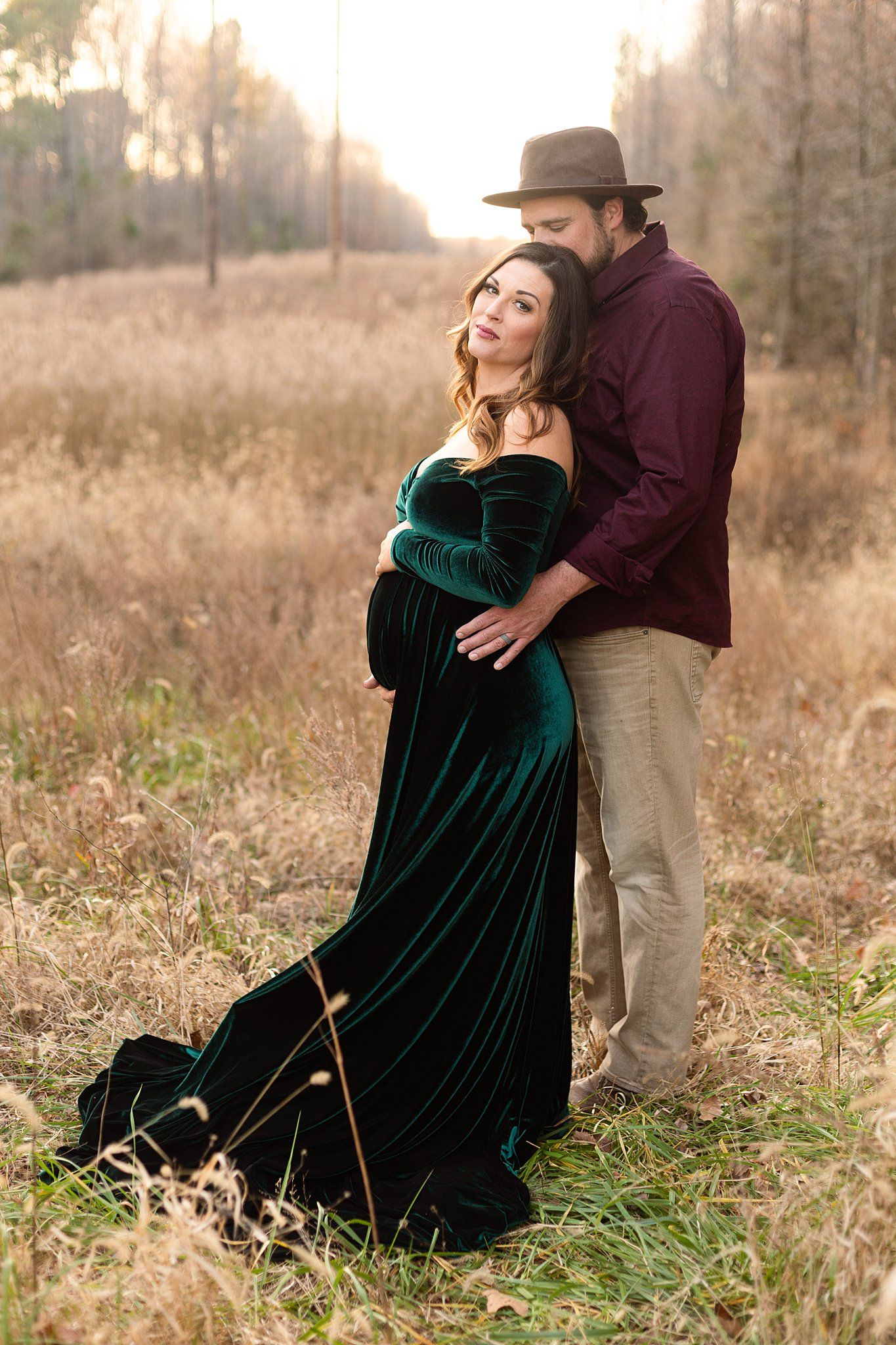 Maternity portrait in golden field featuring couple in burgundy and emerald velvet attire.