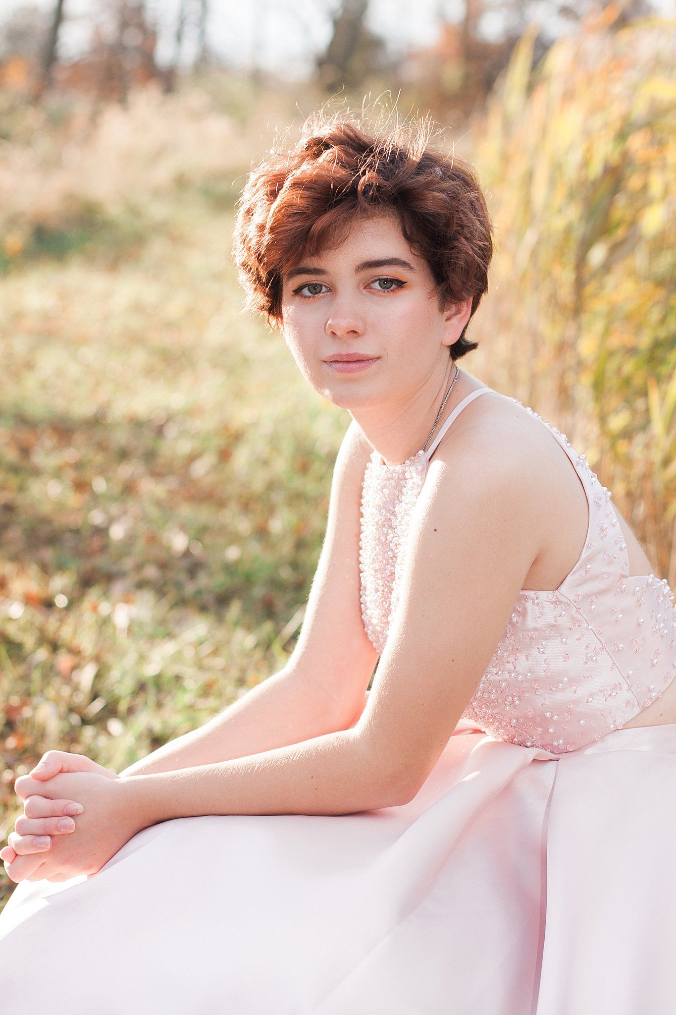 Someone in elegant pink gown sits gracefully among grass at sunset.