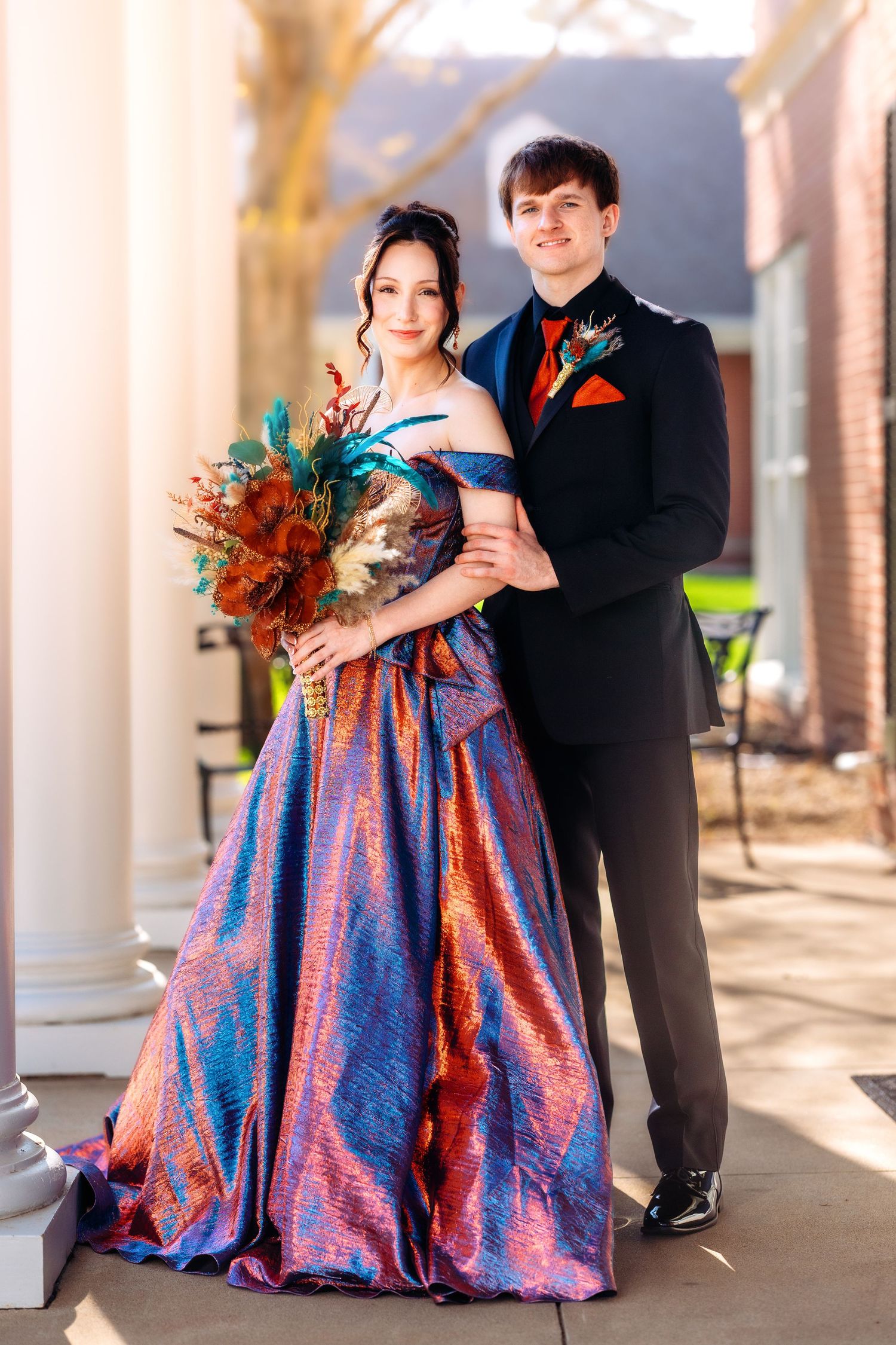 Couple in formal attire poses together at a wedding, with the bride wearing an iridescent blue and red gown.