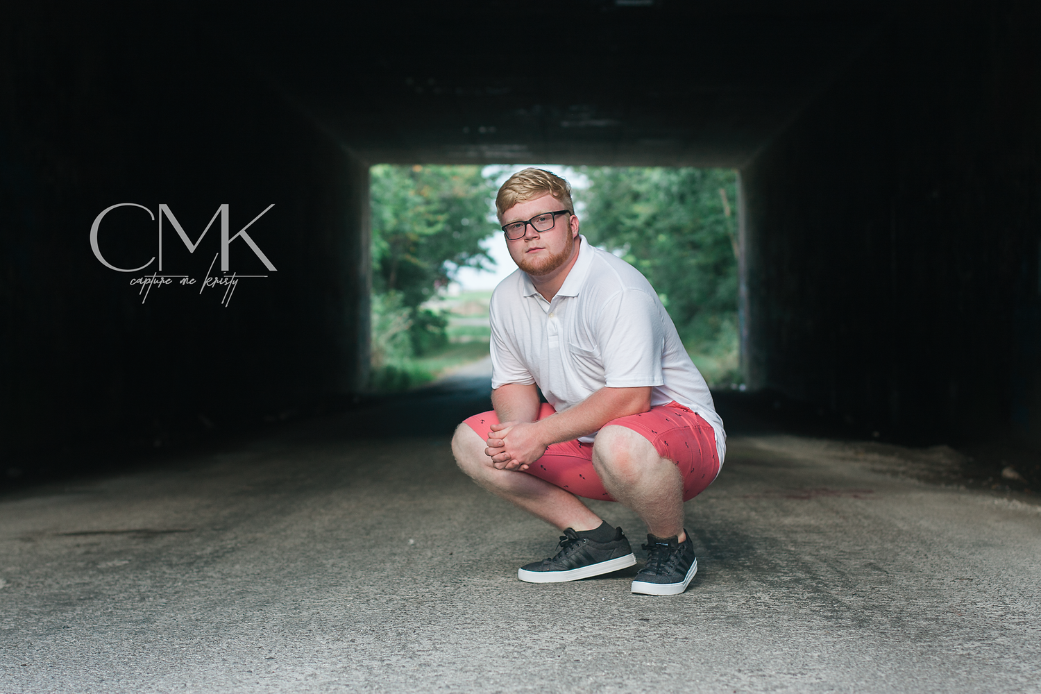 Professional photo of someone in casual wear crouching in a tunnel with green foliage visible in background.