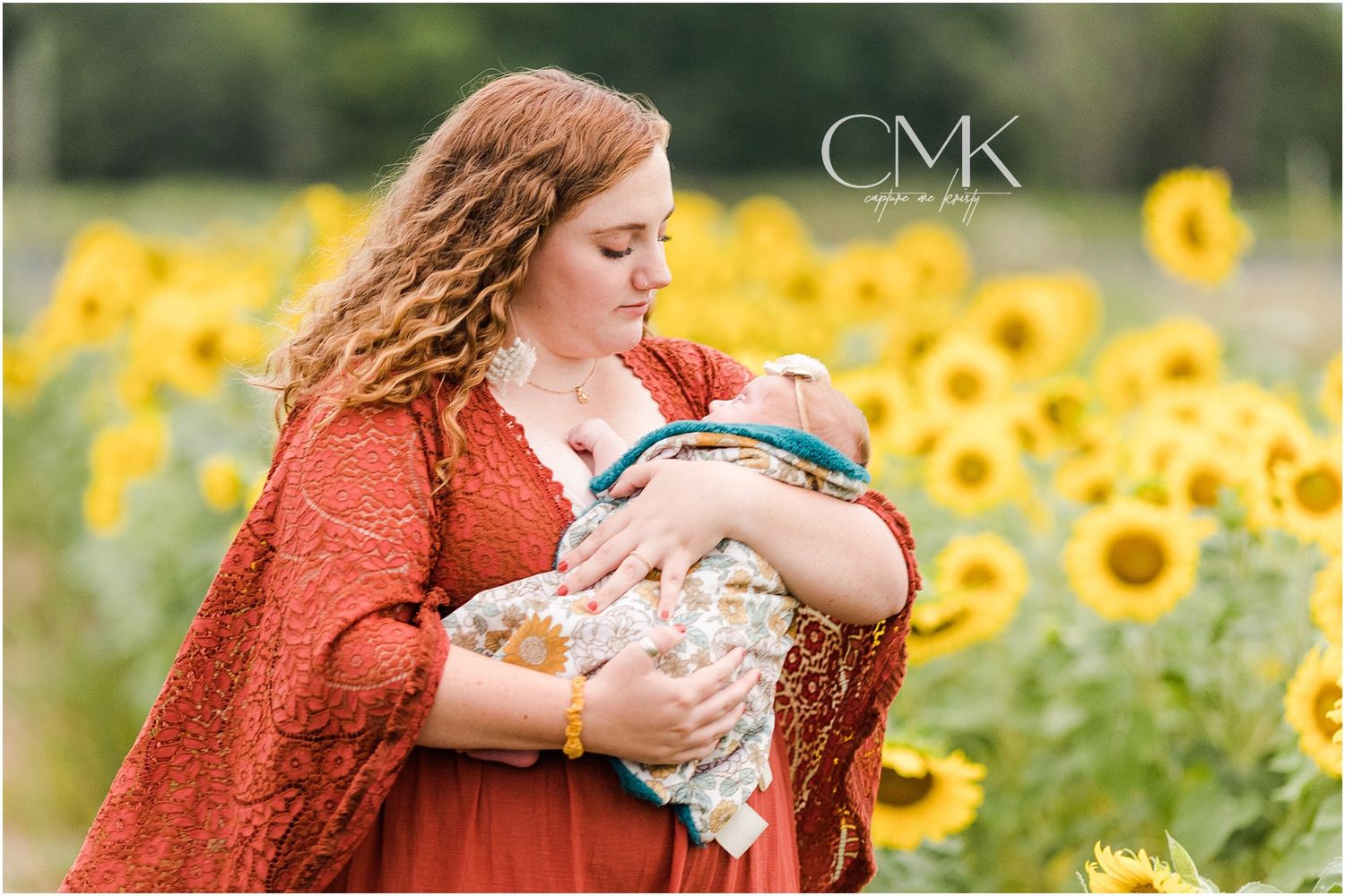Parent in rust dress holding baby wrapped in pattern blanket amidst sunflower field.