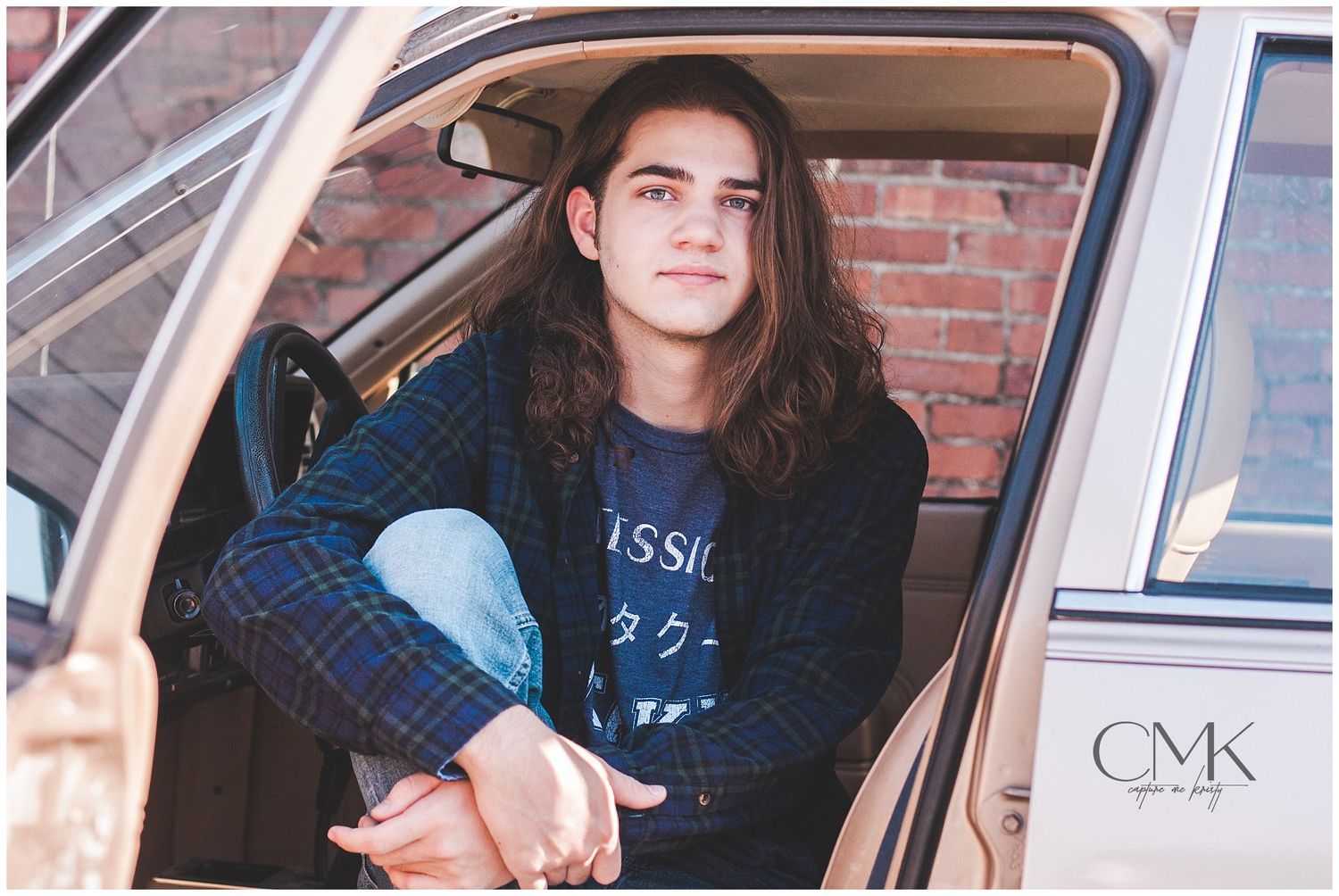 Person in dark shirt sitting in vintage car against brick wall background.