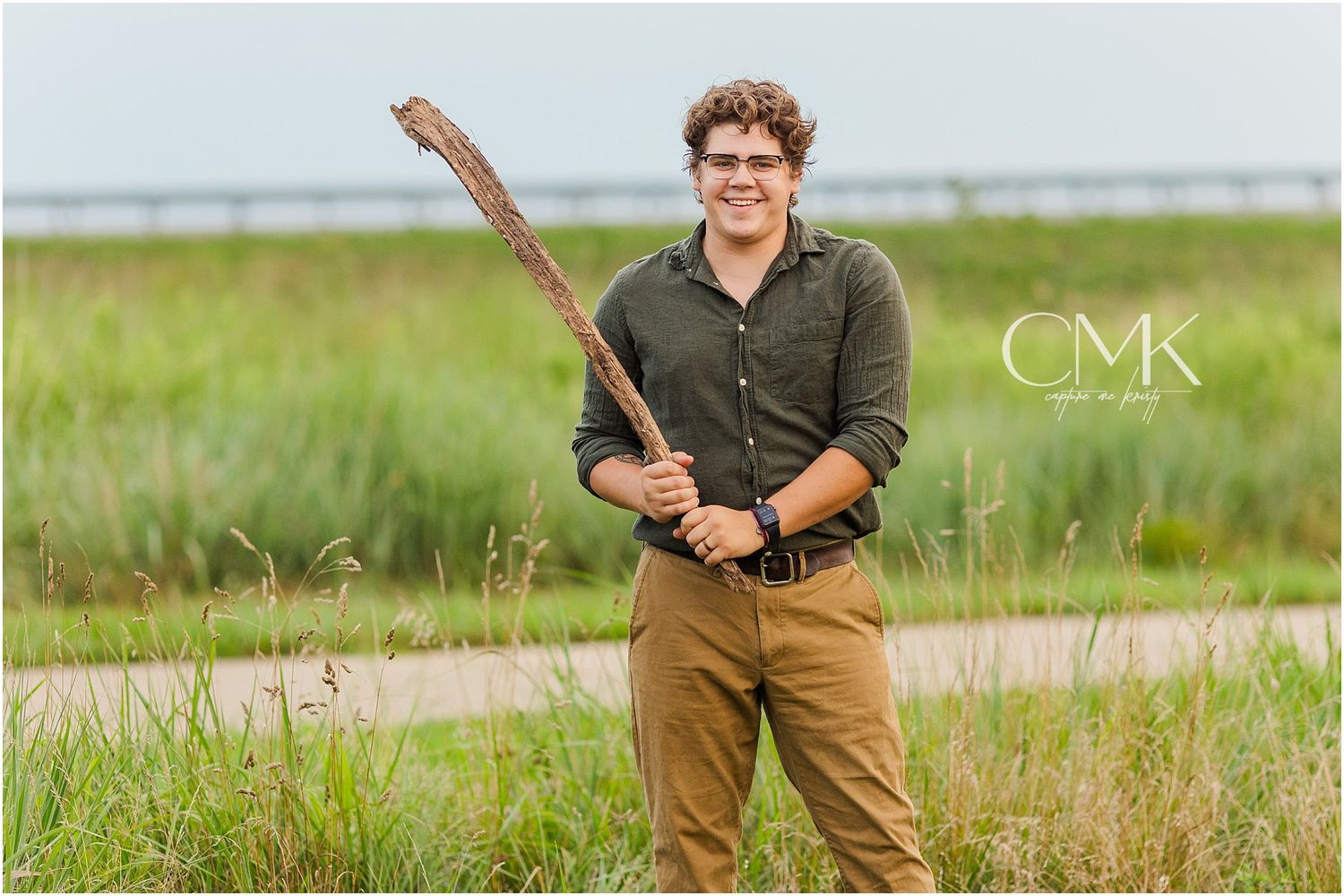 Person in olive shirt and khaki pants holding a large stick while standing in a grassy field.