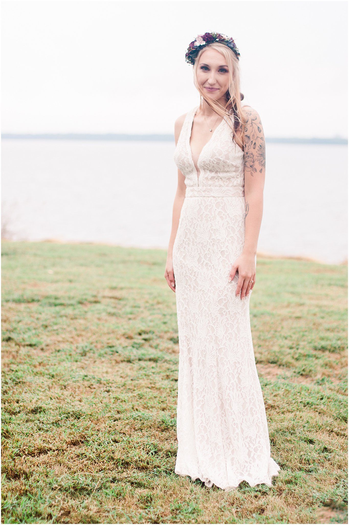 A bride in a lace wedding dress and floral crown poses outdoors against a waterfront backdrop.