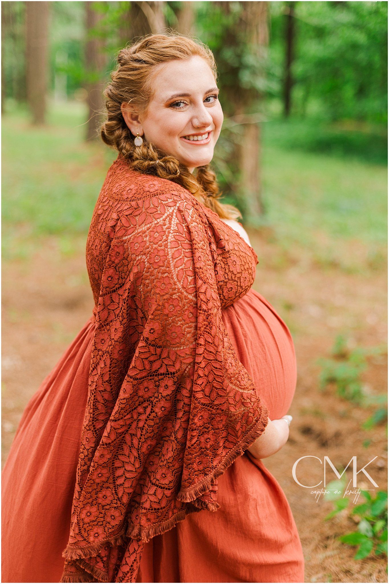 Outdoor maternity portrait in flowing coral lace dress among woodland scenery.
