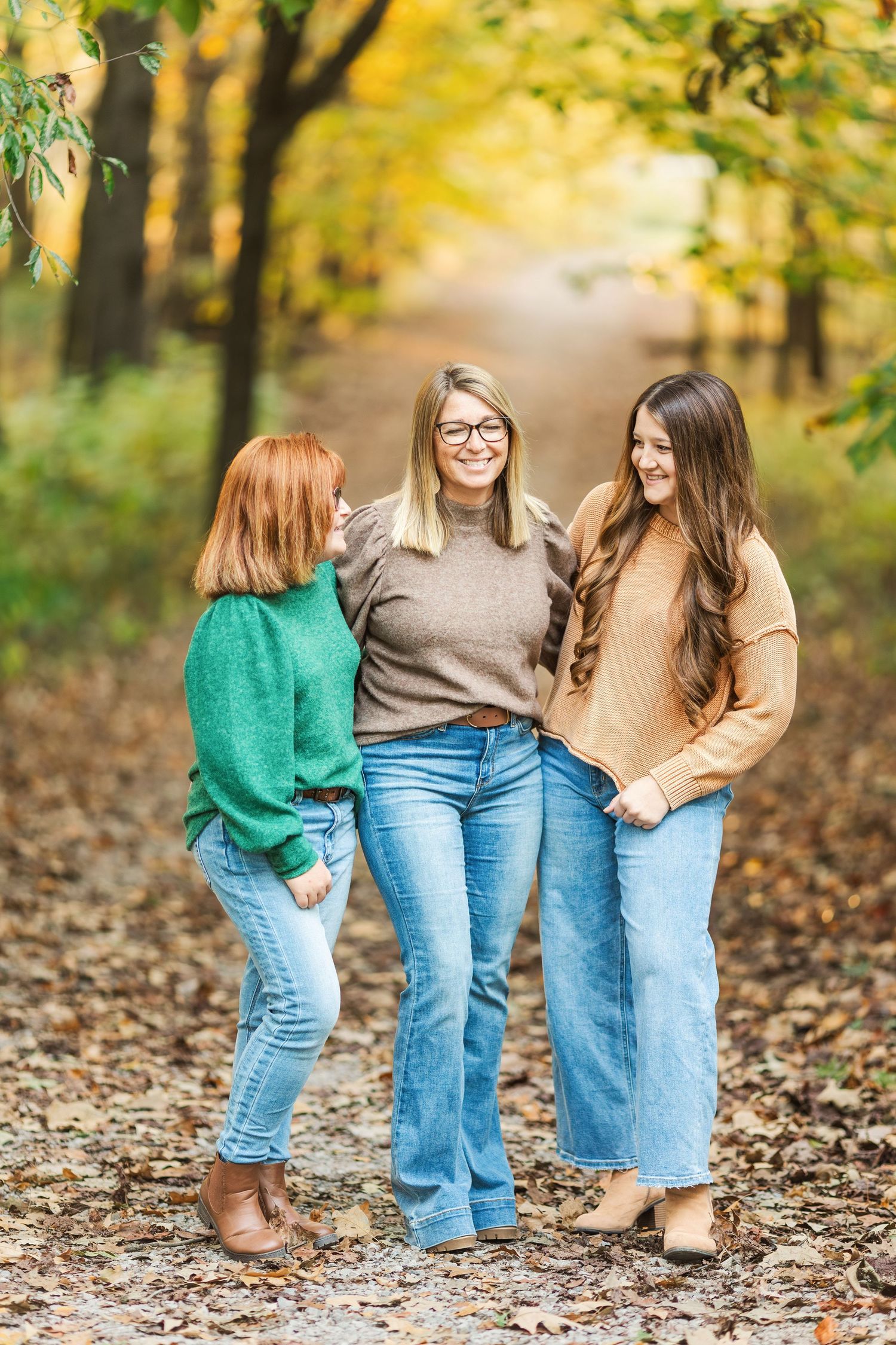 Three friends in casual sweaters and jeans share a moment on a leaf-covered autumn path.