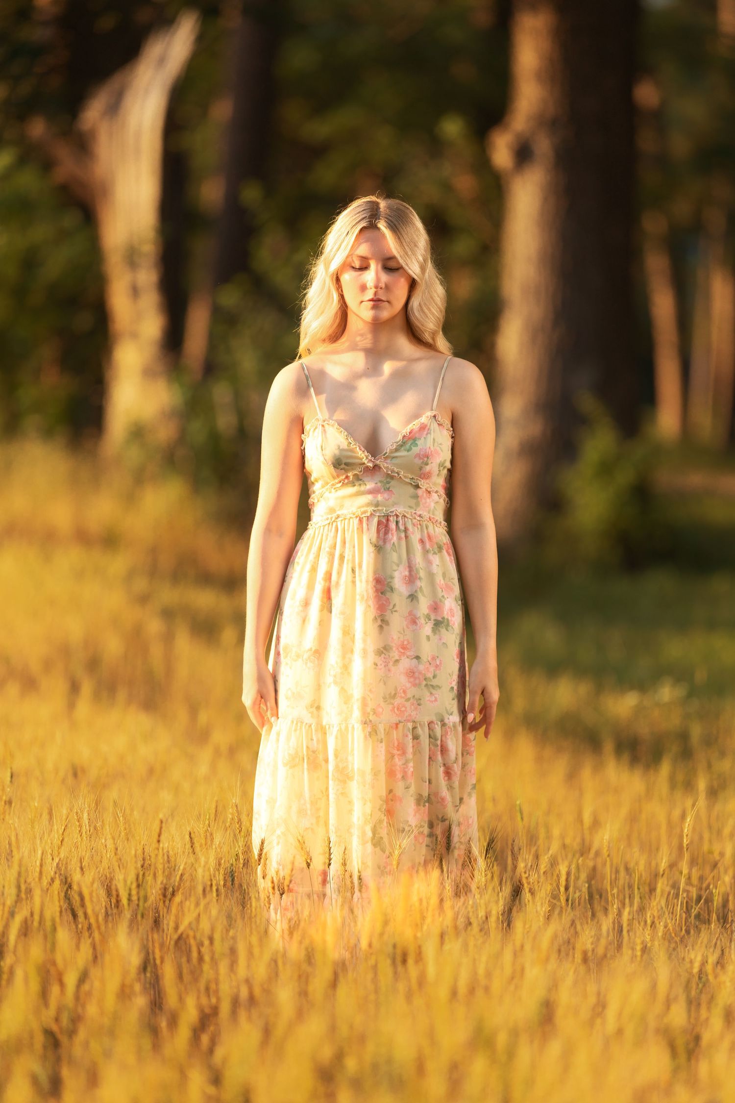 Blonde woman in floral sundress standing in golden wheat field at sunset with trees in background.